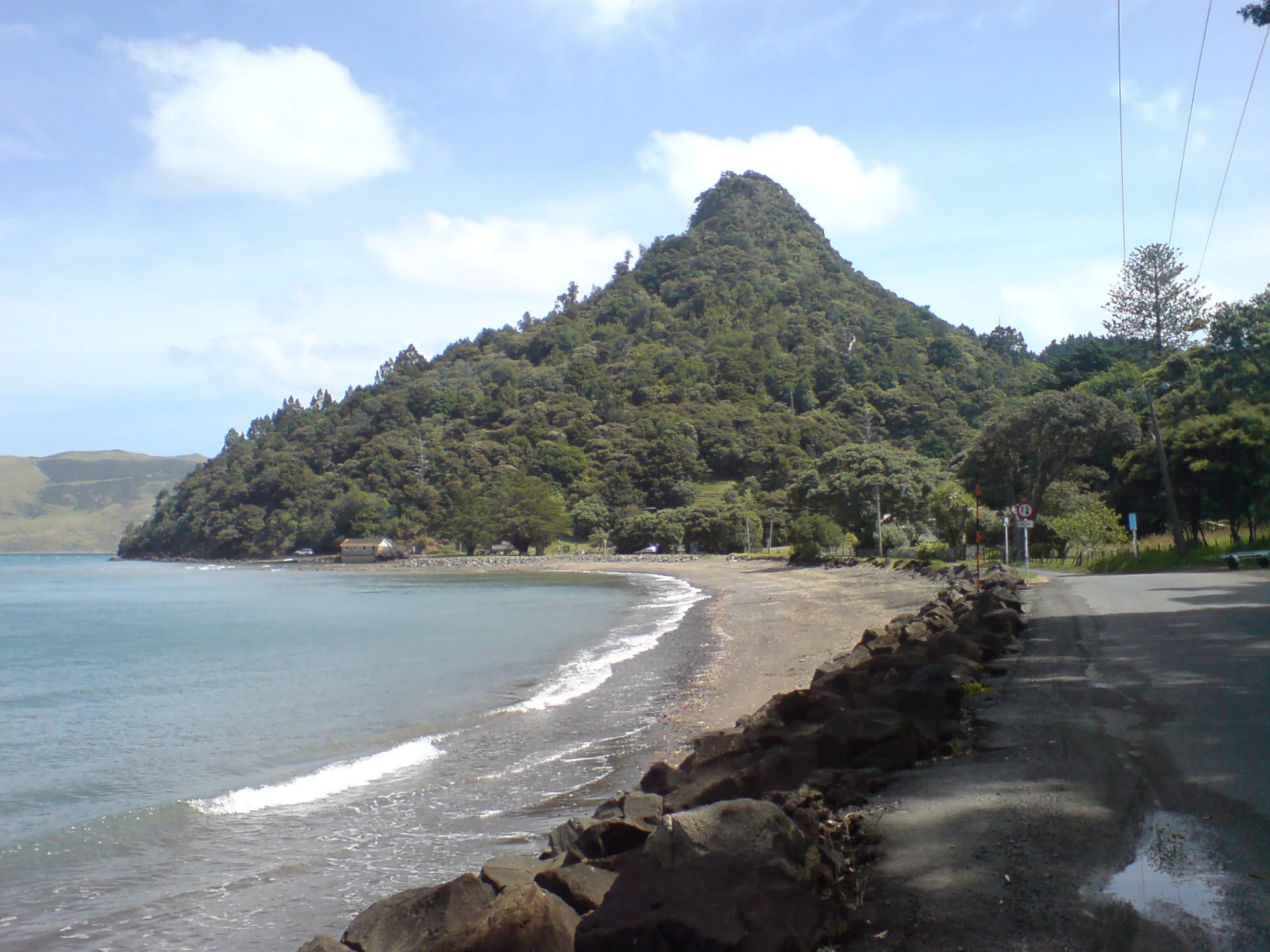 A beach with dark sand curving along the coast, leading toward a prominent forest-covered hill with a road and a low rock wall in the foreground.