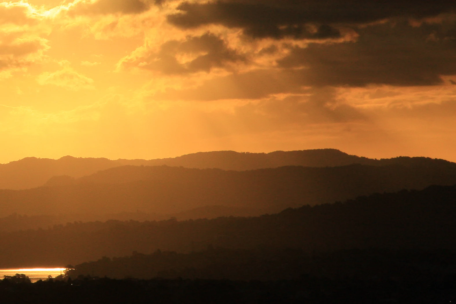 A silhouette of layered mountain ridges against a glowing orange sunset sky with clouds, featuring a glimpse of reflective water in the lower left.