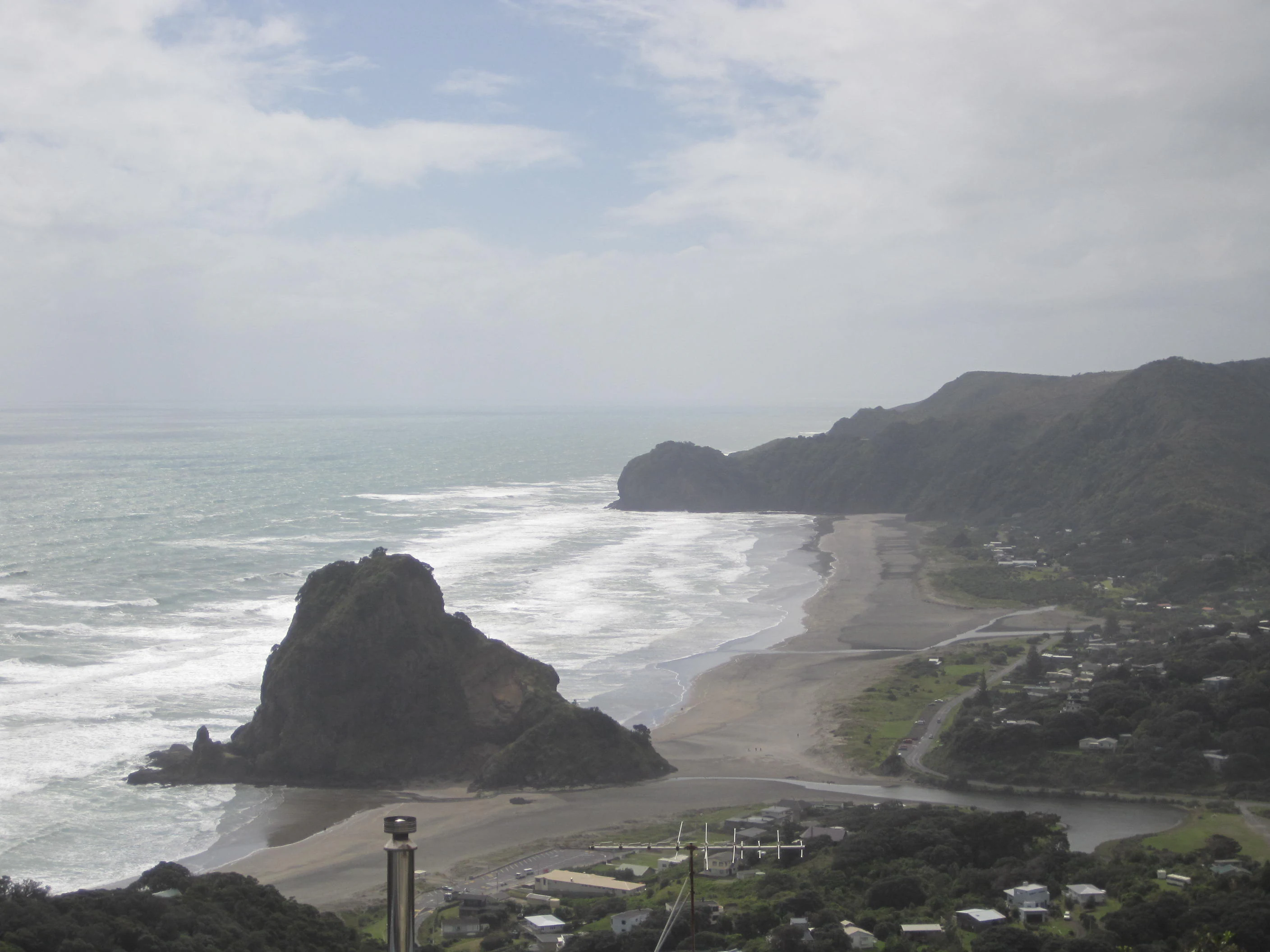 A high-angle view looking down at a large rock formation known as Lion Rock on a sandy beach, with crashing surf and coastal cliffs in the background under a cloudy sky.