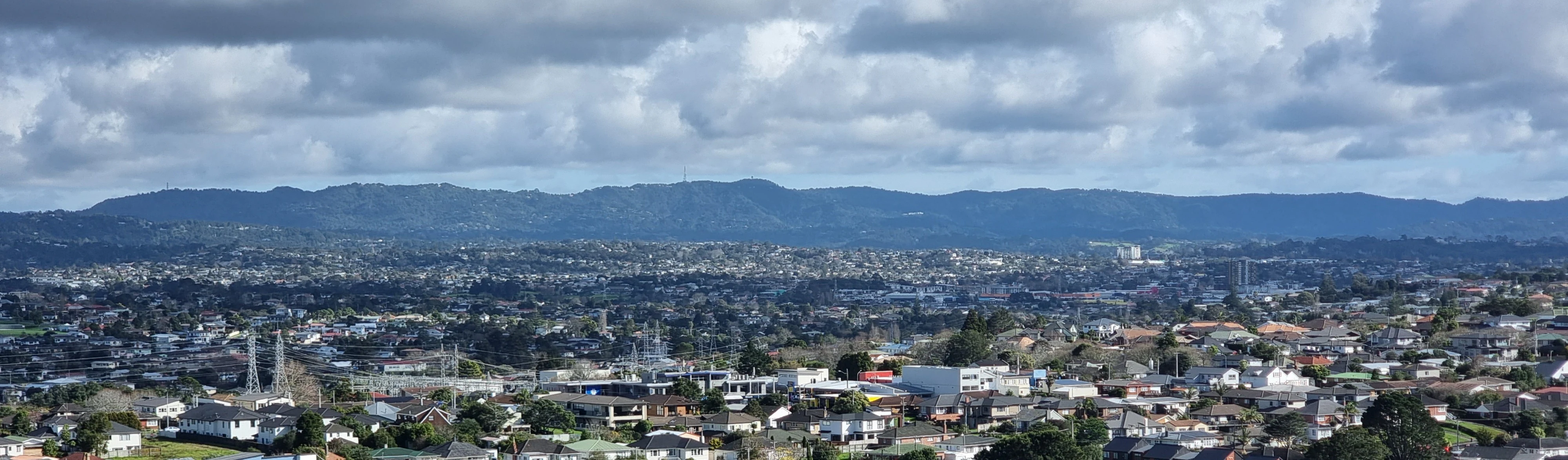 A wide-angle, long-distance view of the forested Waitakere Ranges stretching across the horizon, with suburban buildings and infrastructure in the foreground and midground.