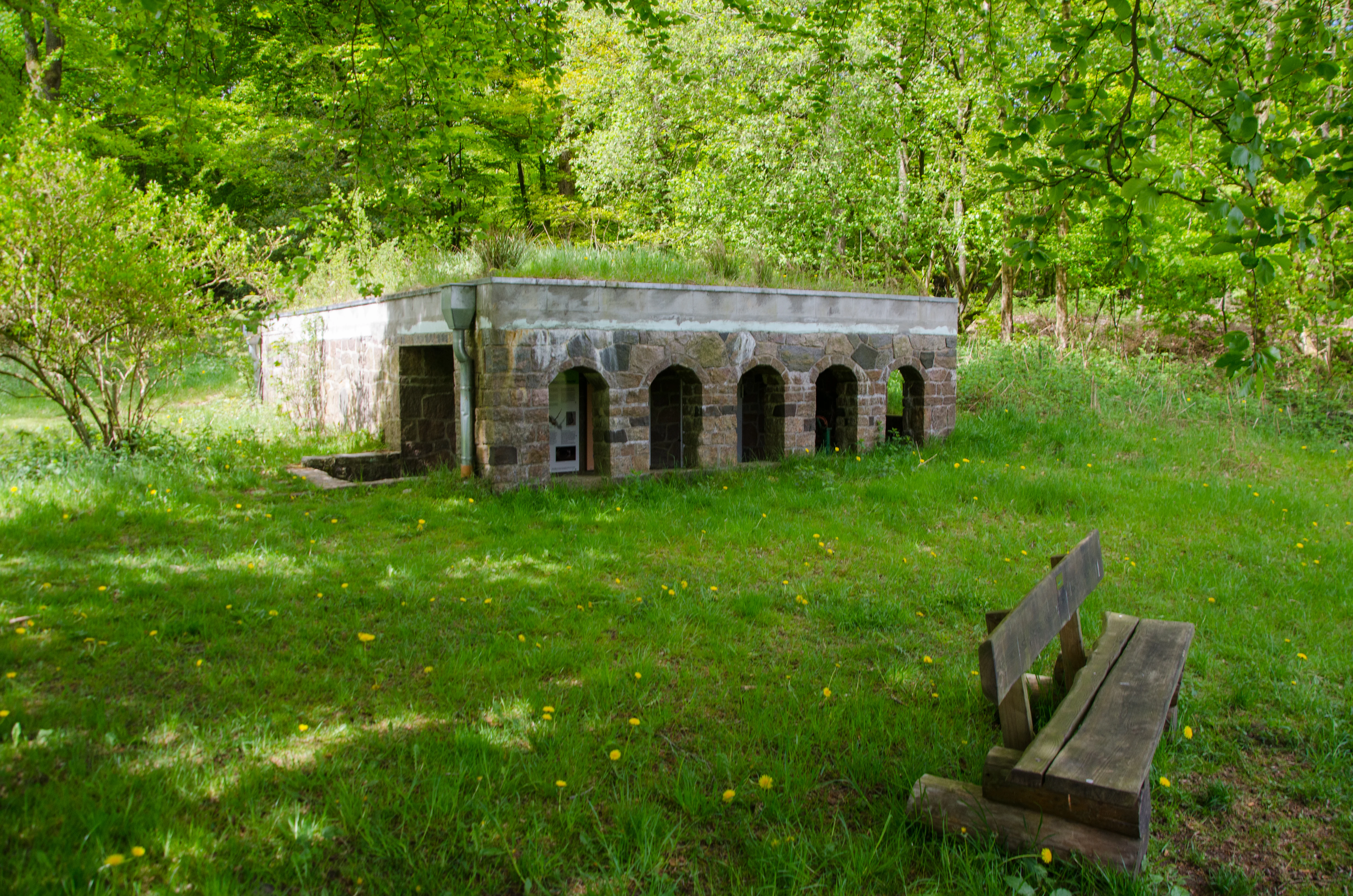 A low, stone-walled structure with multiple arched openings, partially covered by earth and grass, located in a woodland clearing with a wooden bench in the foreground.