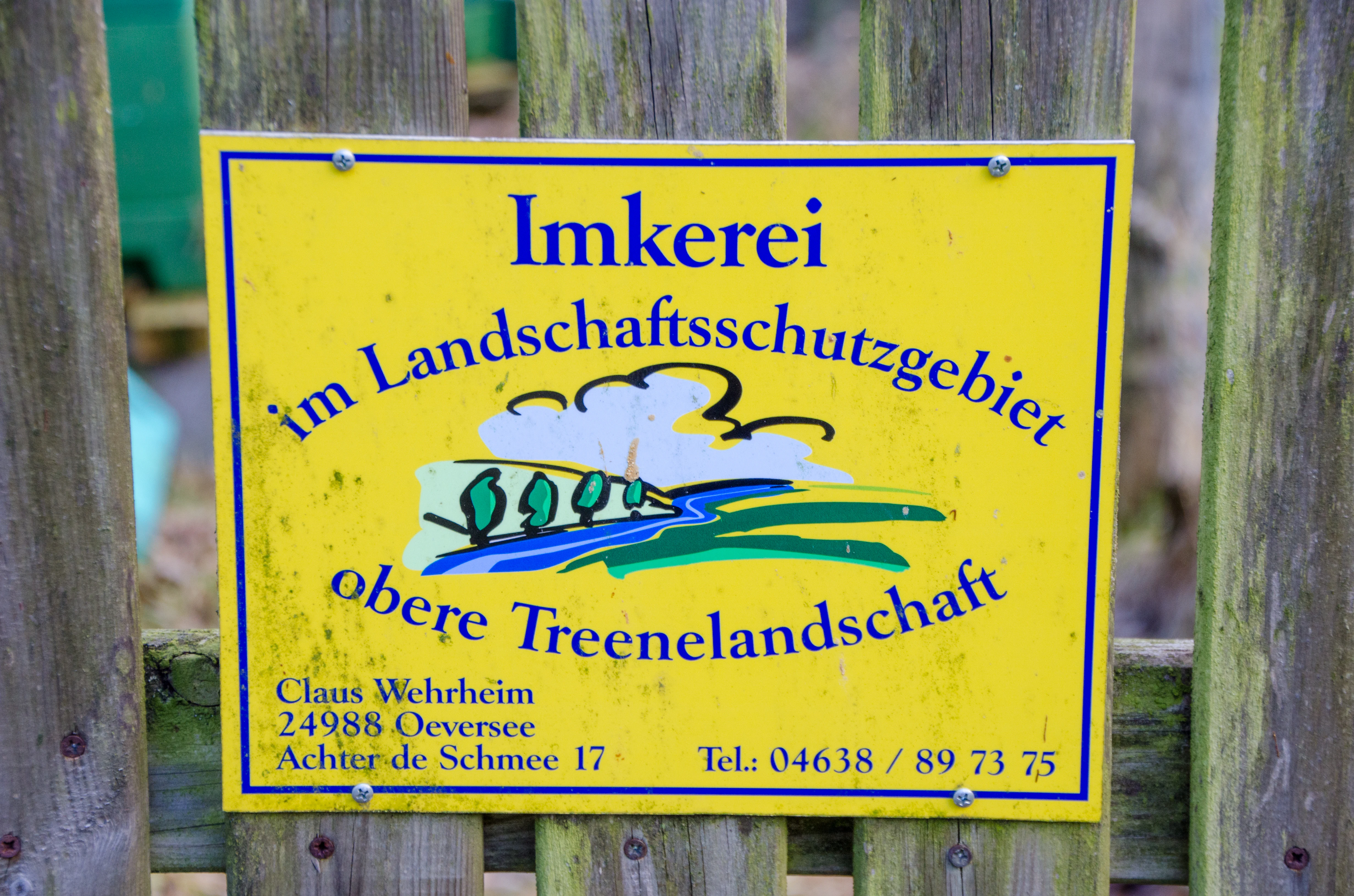 A yellow rectangular sign attached to a wooden fence reads Imkerei im Landschaftsschutzgebiet obere Treenelandschaft with contact details below, featuring a small illustration of a landscape with a river.