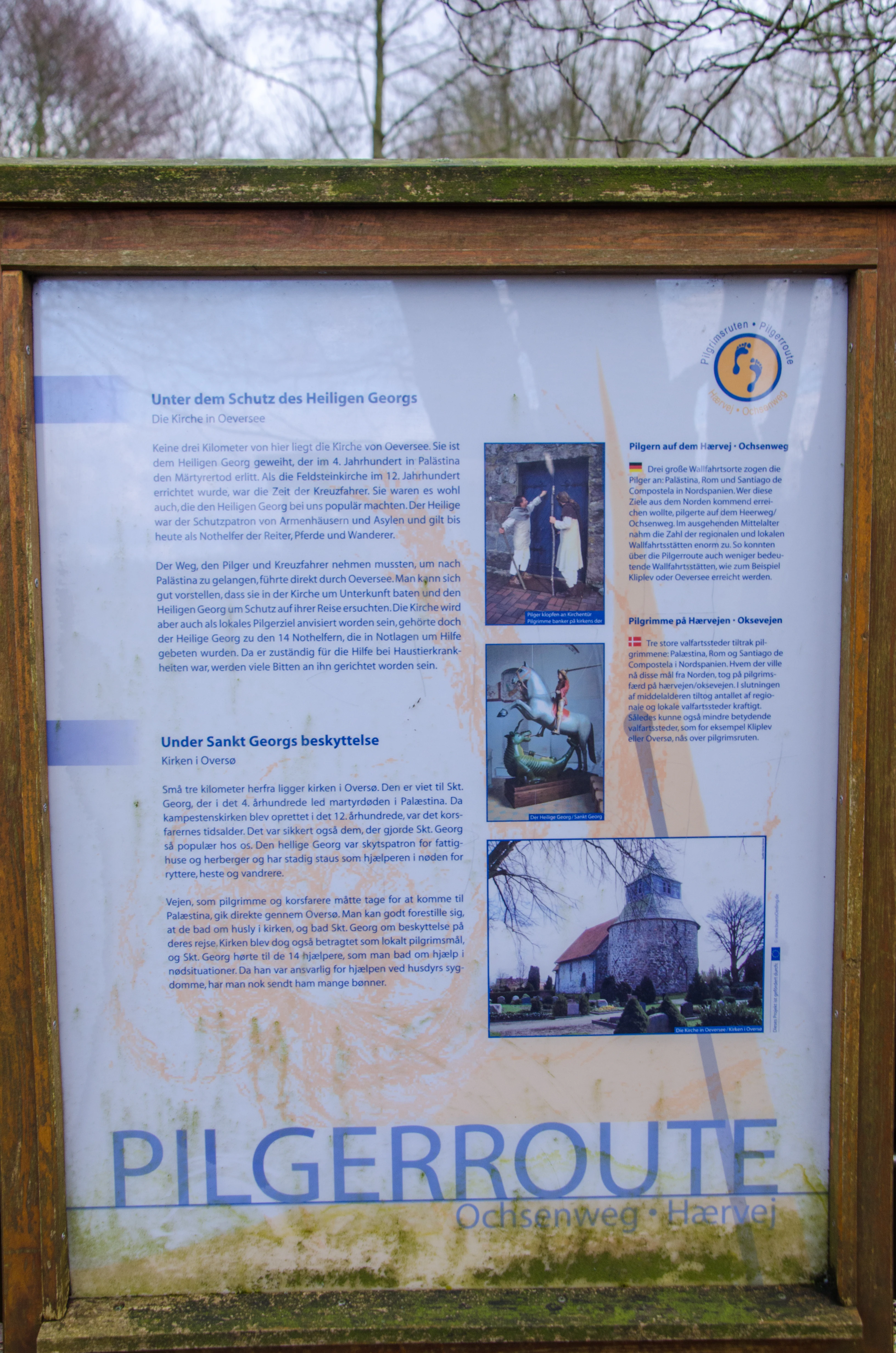 A wooden-framed information board with text and images in German and Danish about the 'Pilgerroute' or pilgrim path, featuring small photos of a church and historical imagery.