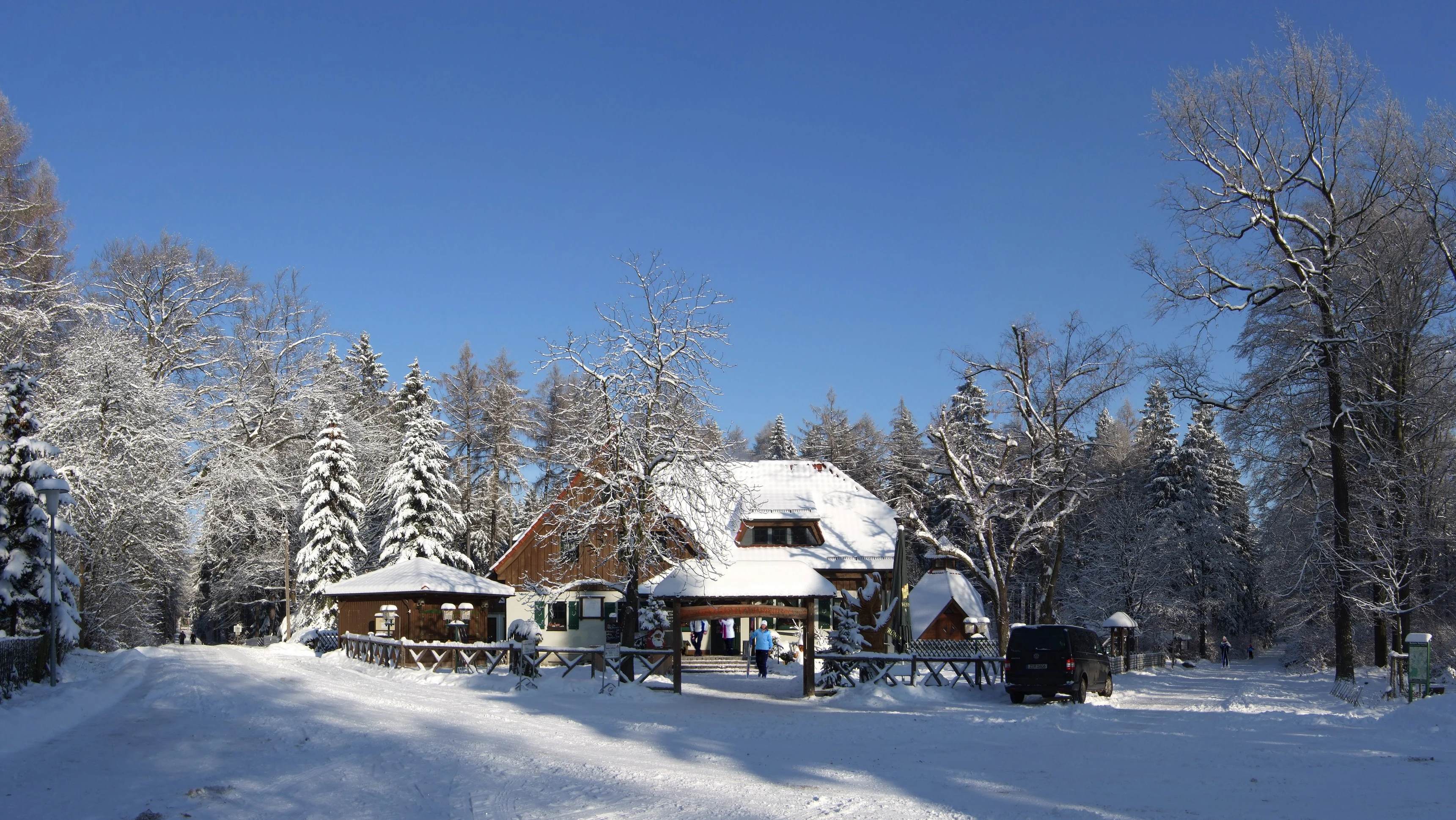 A snow-covered building with brown walls and a gabled roof in the middle of a forest during winter, with snow-covered trees and a cleared path in the foreground.