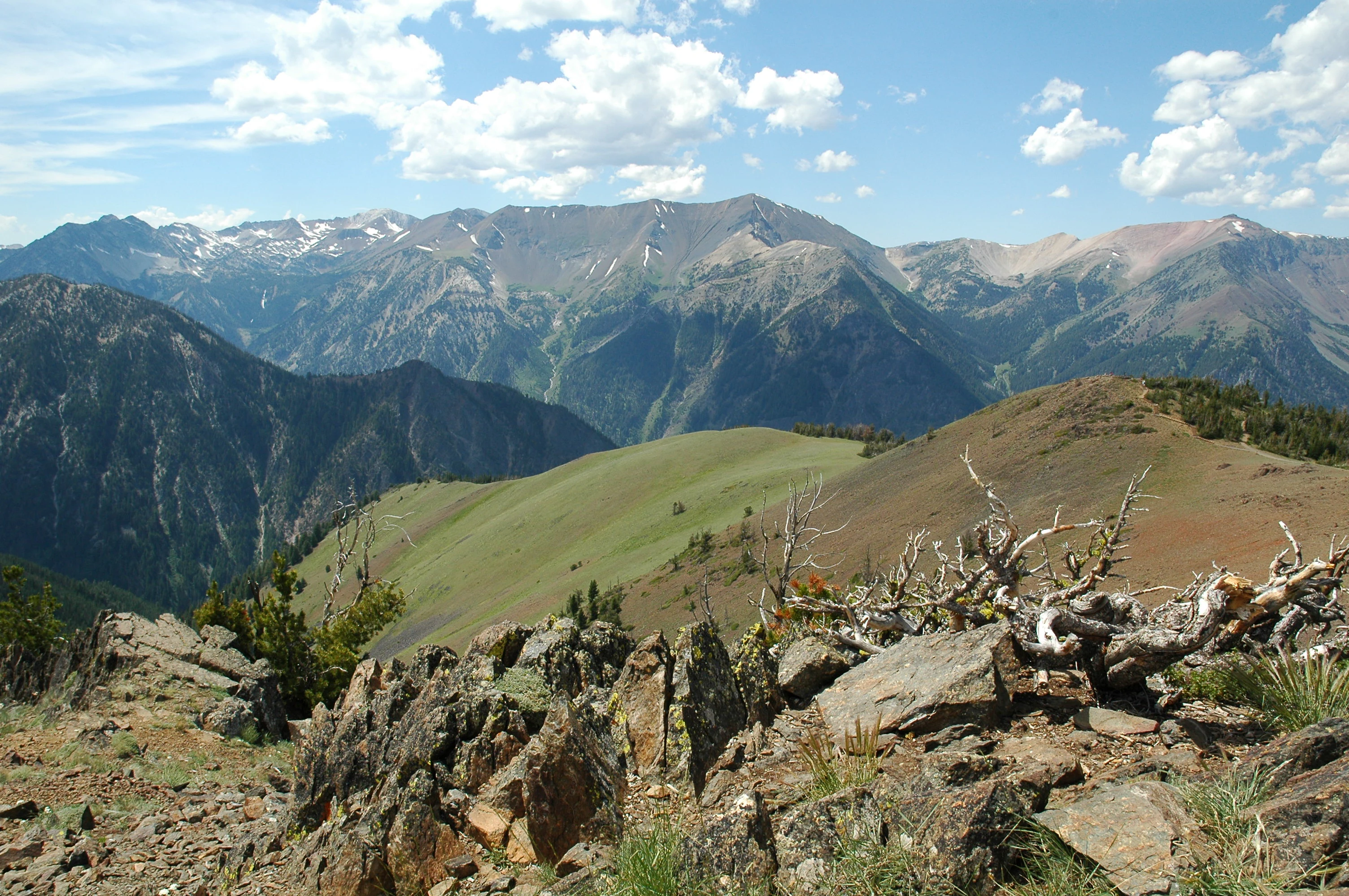 A wide, eye-level mountain landscape showing a series of jagged, rocky peaks against a blue sky with light clouds, looking over a foreground of rocky soil and a dried, weathered tree stump.
