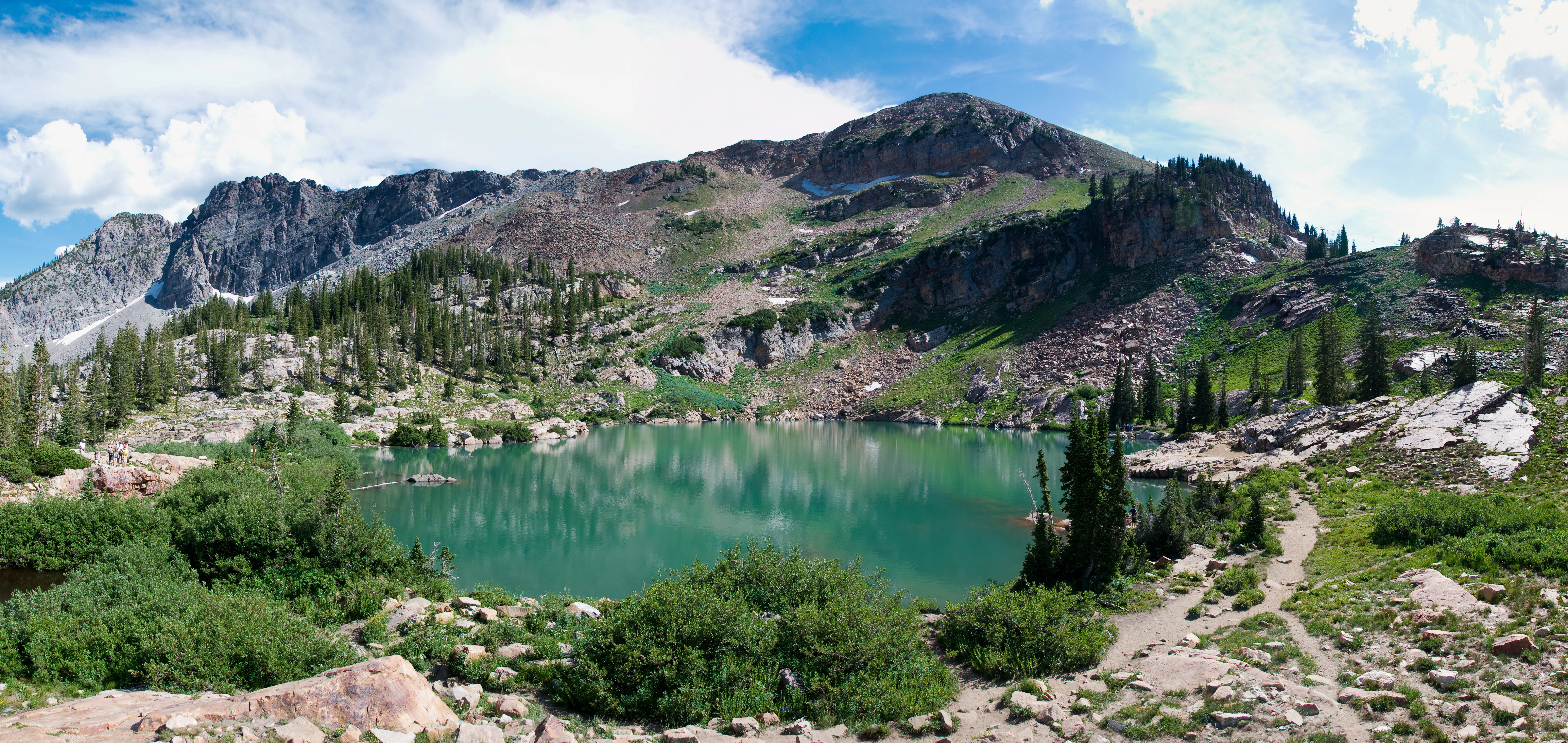 Vista panorámica de un lago alpino turquesa rodeado por una gran ladera montañosa con pinos dispersos y terreno rocoso.