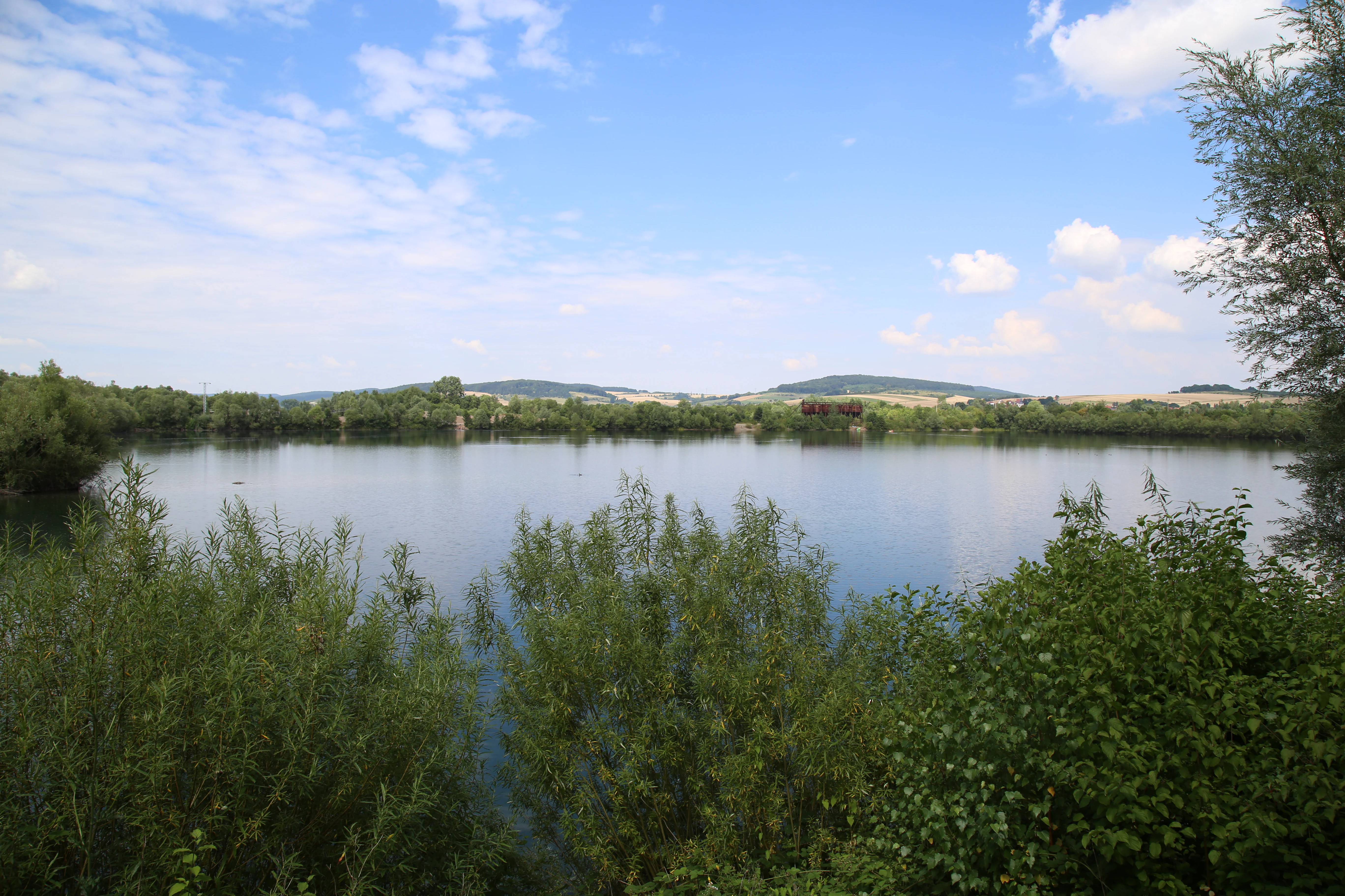 A wide view of a calm lake surrounded by lush green trees and shrubs under a blue sky with light clouds, with rolling hills visible in the background.
