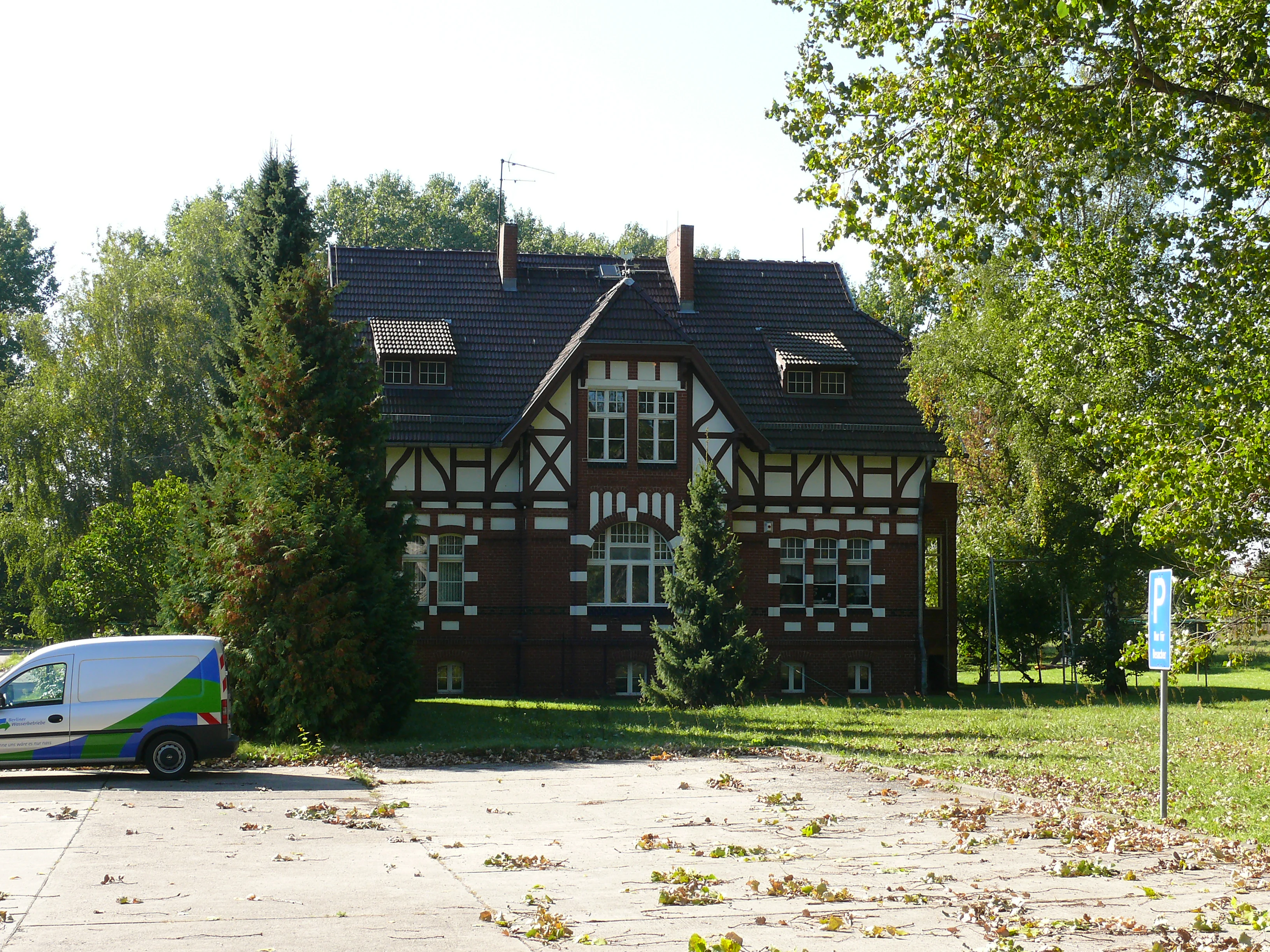 A historic, timber-framed brick building stands in a grassy park setting, with a white utility van parked in the foreground on a concrete area.