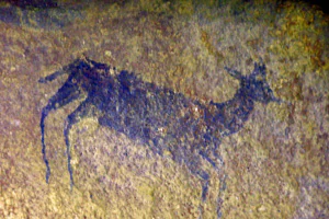 An ancient rock painting in dark pigment on a textured, sandy-colored rock surface showing the silhouette of a four-legged animal resembling a small antelope.