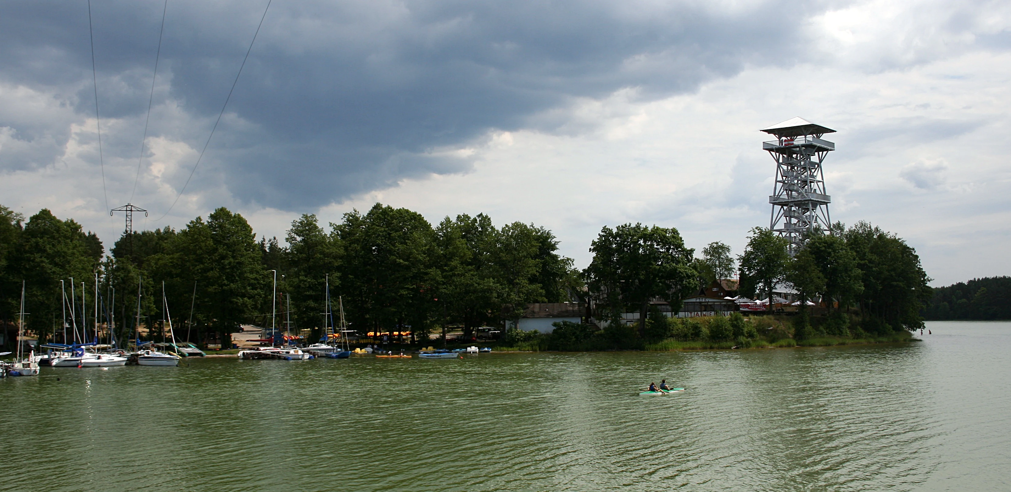 A metal observation tower rises above a tree line on the shore of a large lake, with several sailboats moored nearby under a cloudy sky.