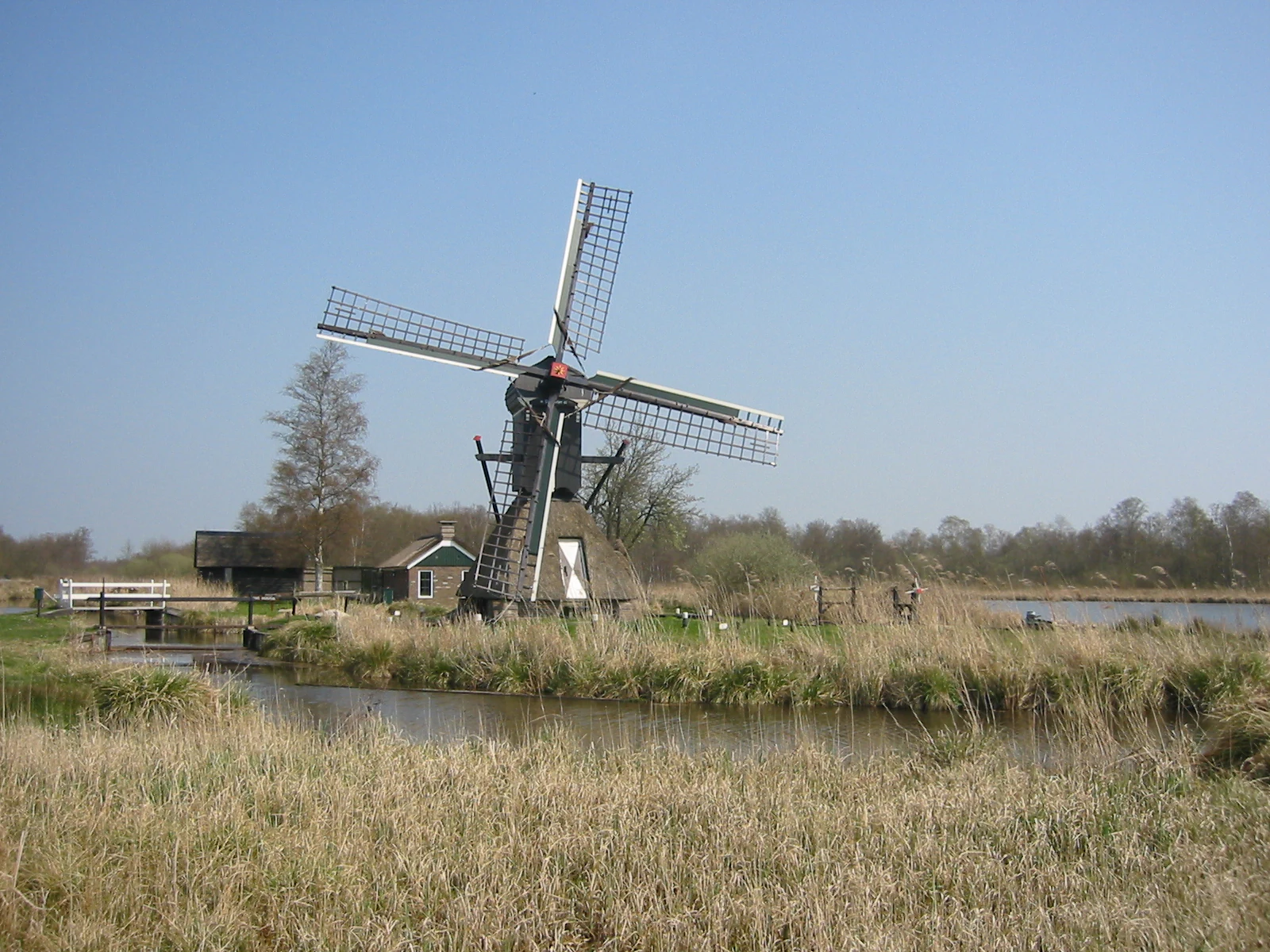A wooden windmill with four blades stands near a body of water in a grassy field. The sky is clear and blue with some trees in the background.