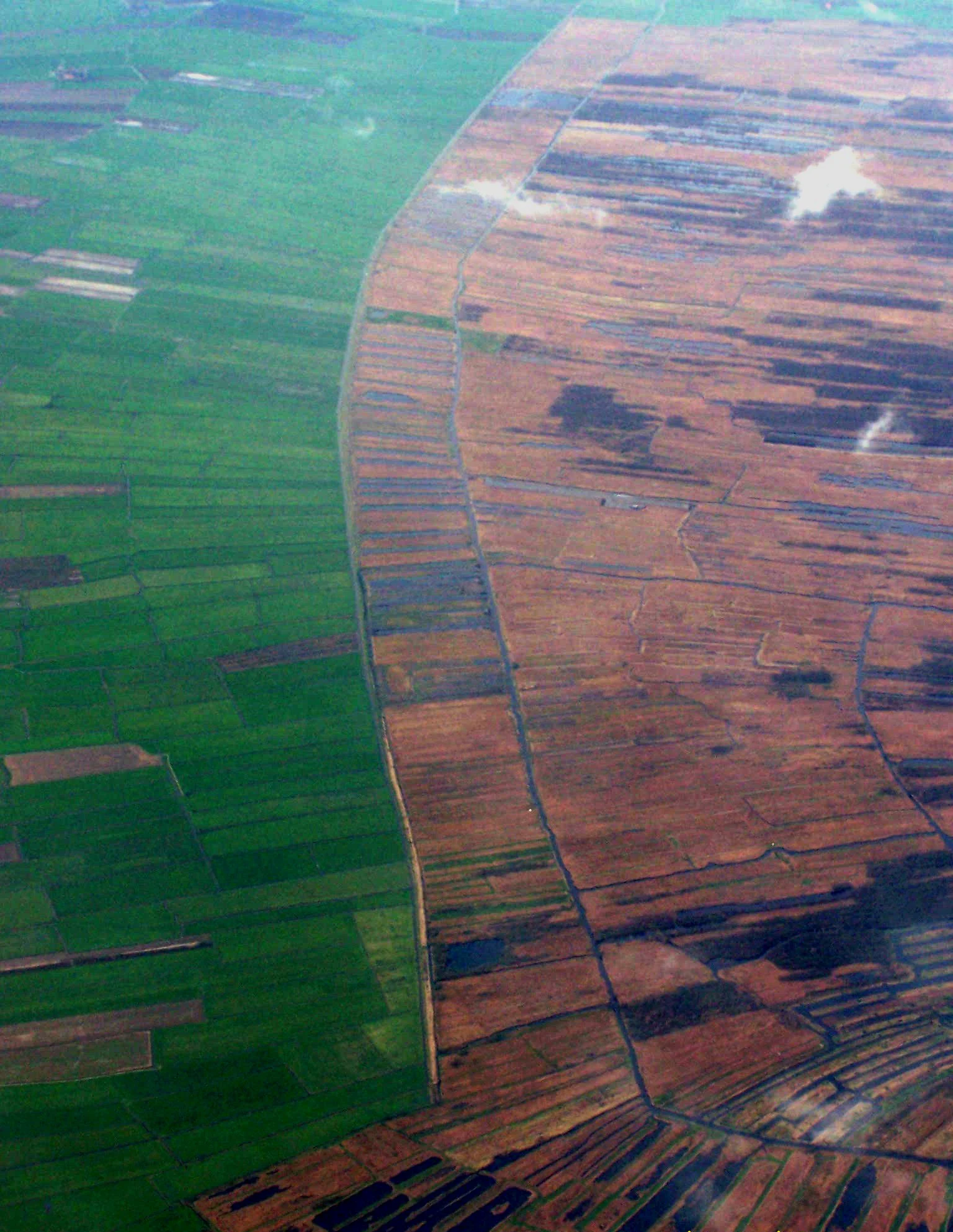 Aerial view of patchwork fields with varying shades of green and brown, some water patches visible, and distinct boundaries between agricultural plots.