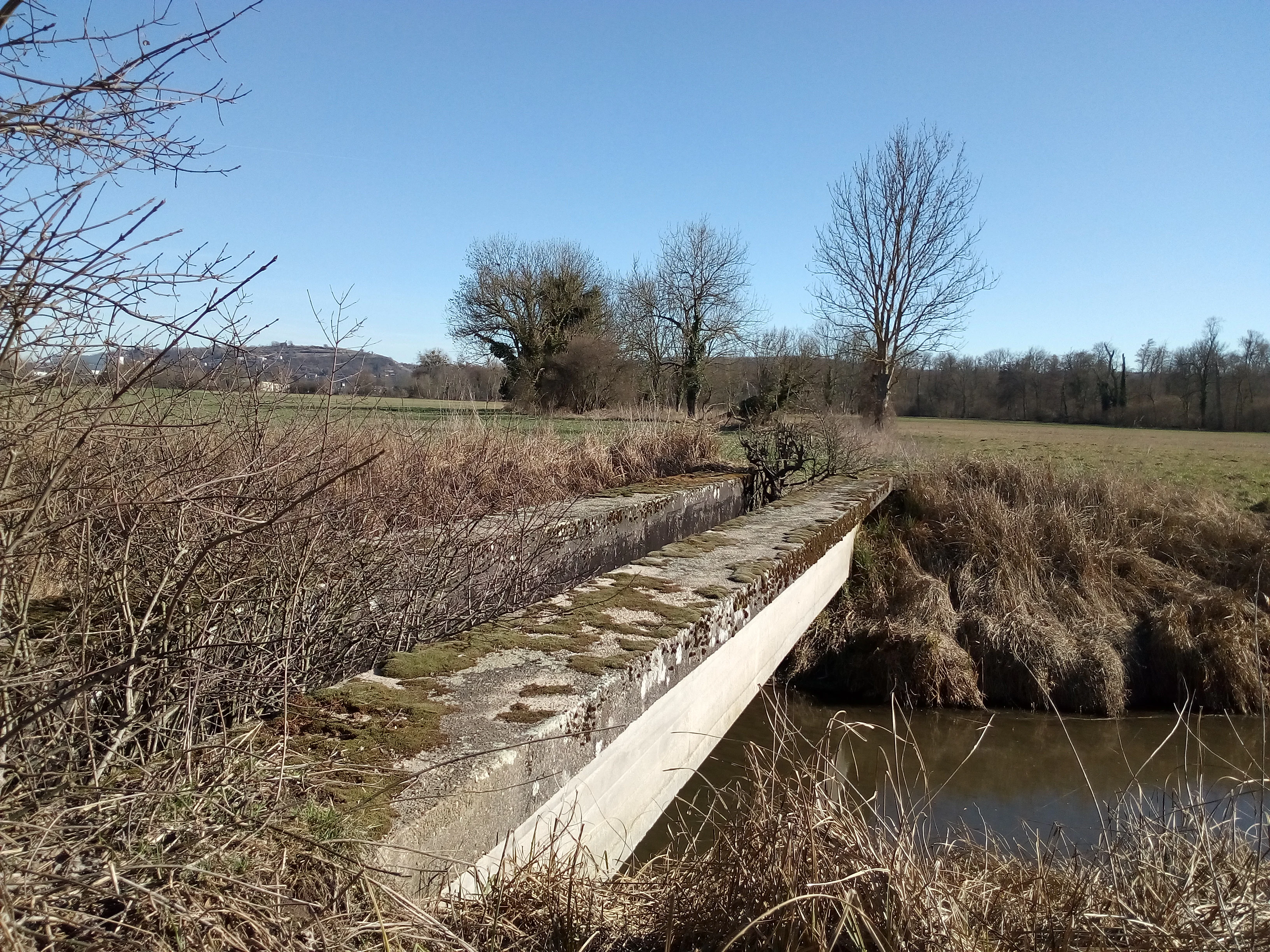 A weathered, moss-covered concrete aqueduct bridge crossing a small stream in a meadow landscape under a clear blue sky.