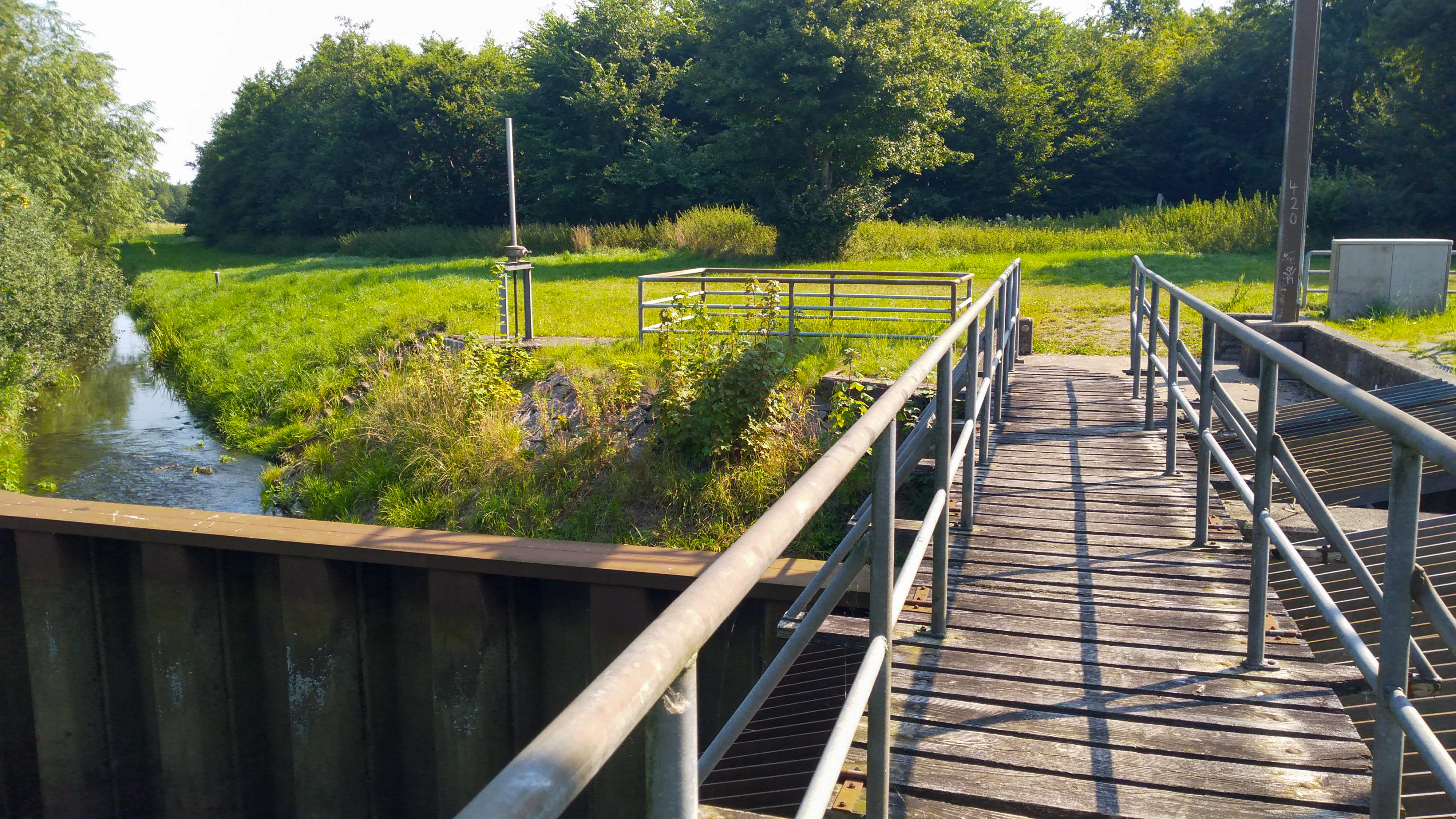 A wooden pedestrian bridge with metal railings spans a stream in a lush, green landscape with trees and grass.