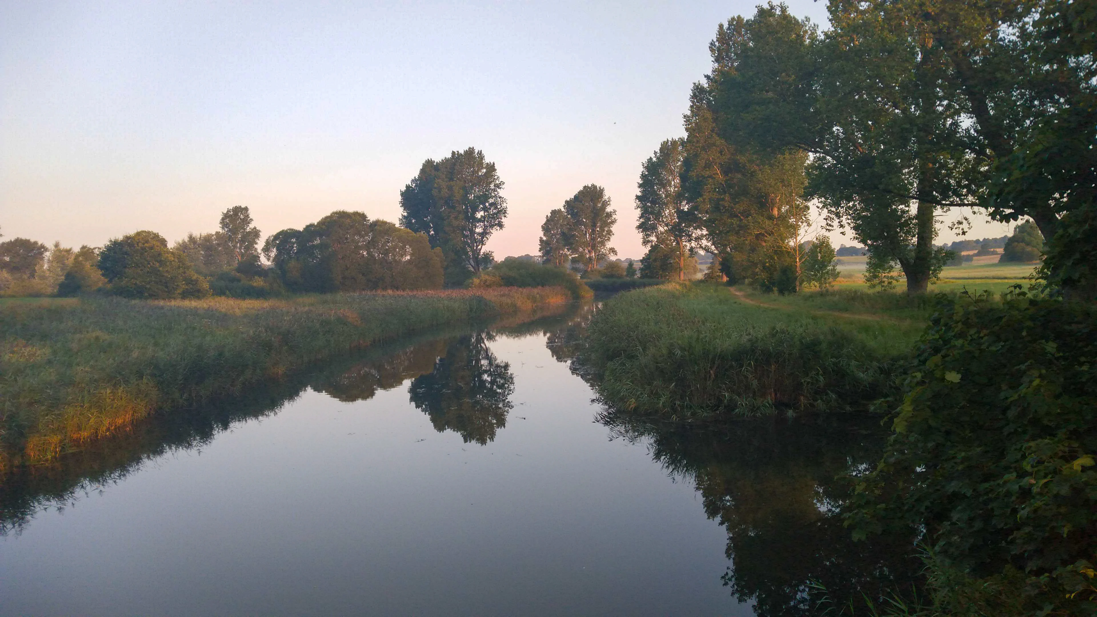 A calm, reflective stream flows through a lush, green landscape under a soft, light-filled sky.
