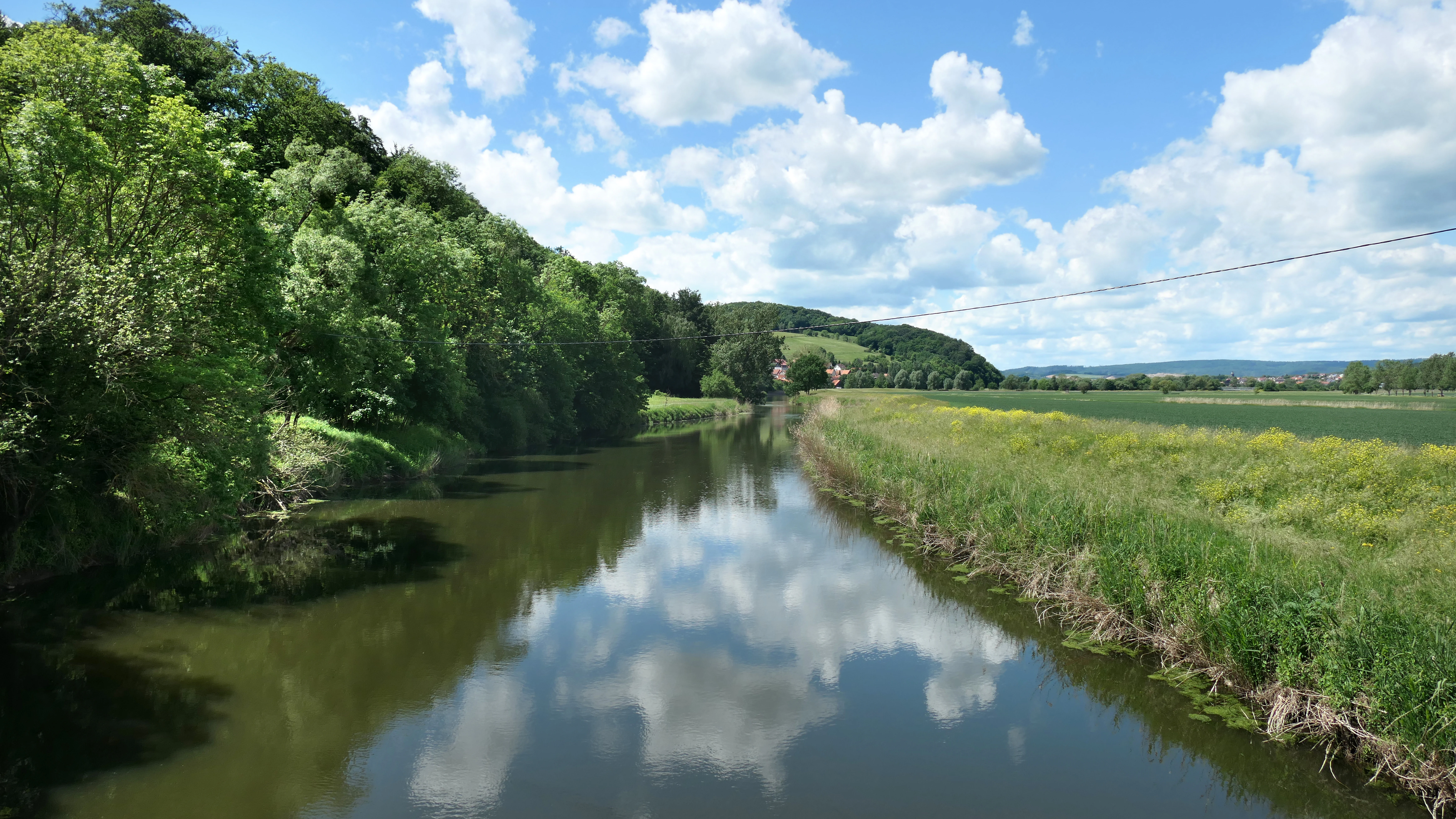 A calm, reflective river stretching into the distance, lined by dense green trees on one bank and an open grassy field on the other under a bright blue sky with scattered clouds.