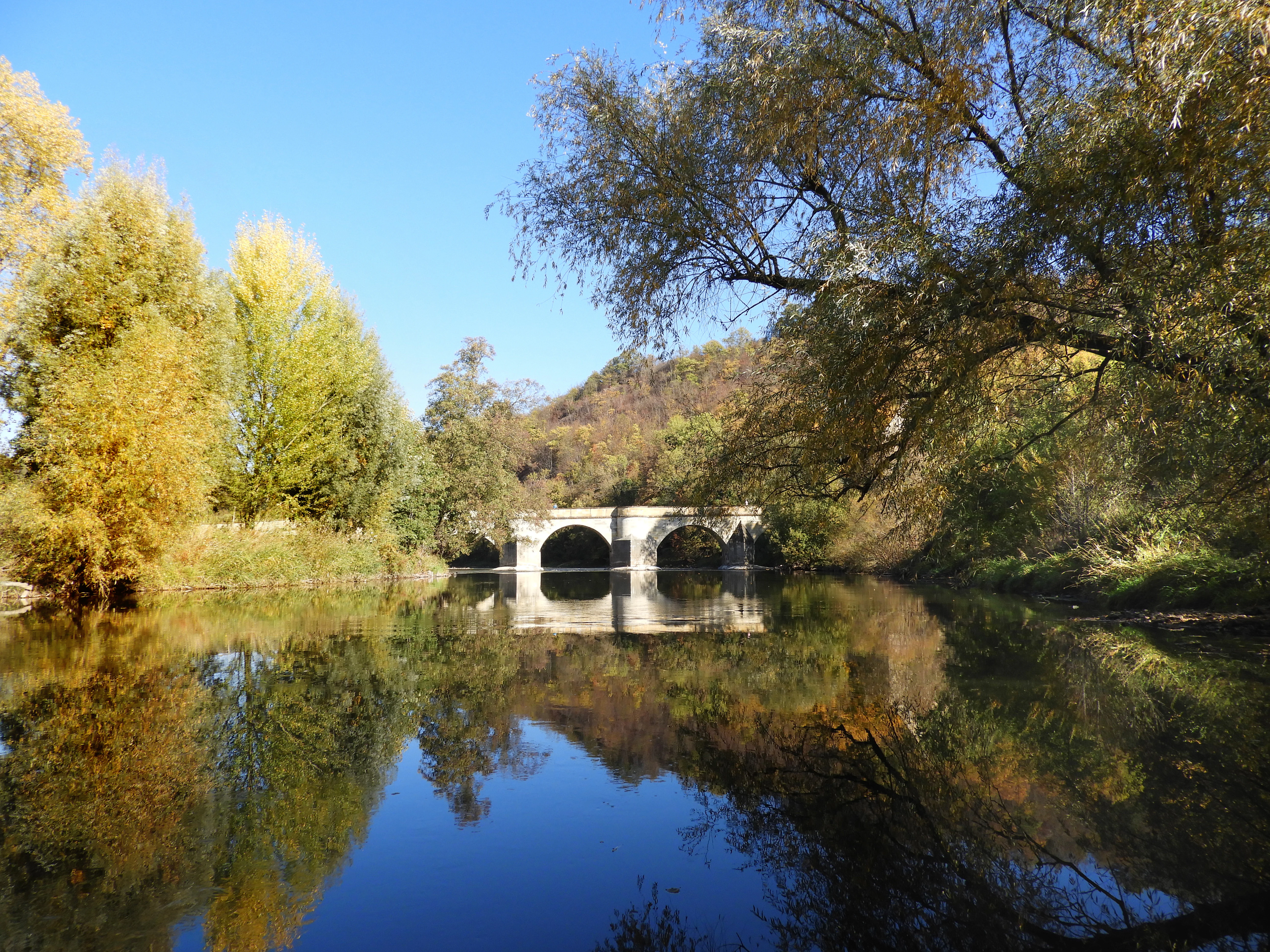 A calm, reflective Werra river flows under a stone arch bridge in Creuzburg, surrounded by deciduous trees with yellow and orange autumn foliage under a clear blue sky.
