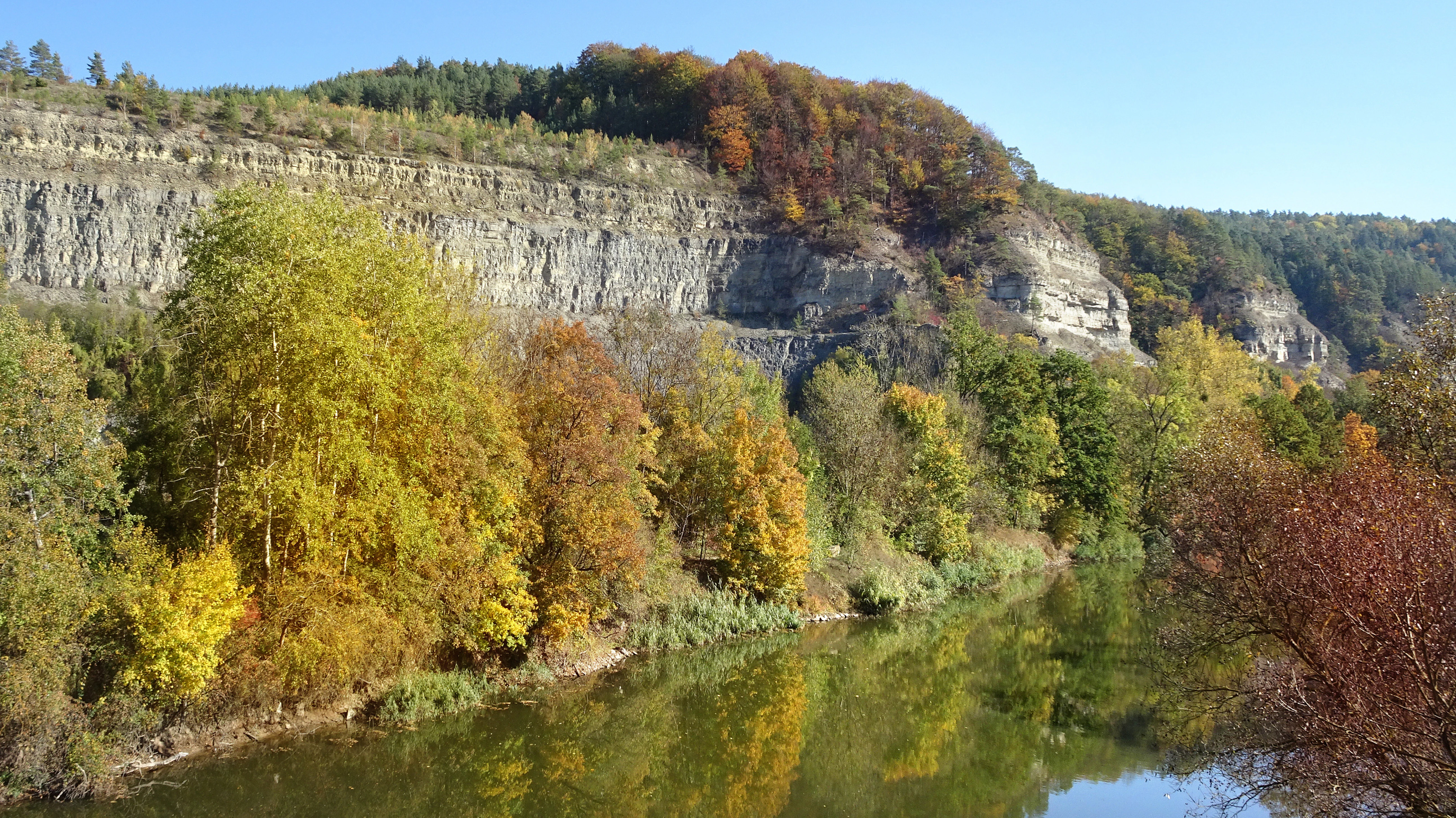 A wide view of a river reflecting the sky, framed by trees with autumn foliage and a large limestone cliff face in the background.