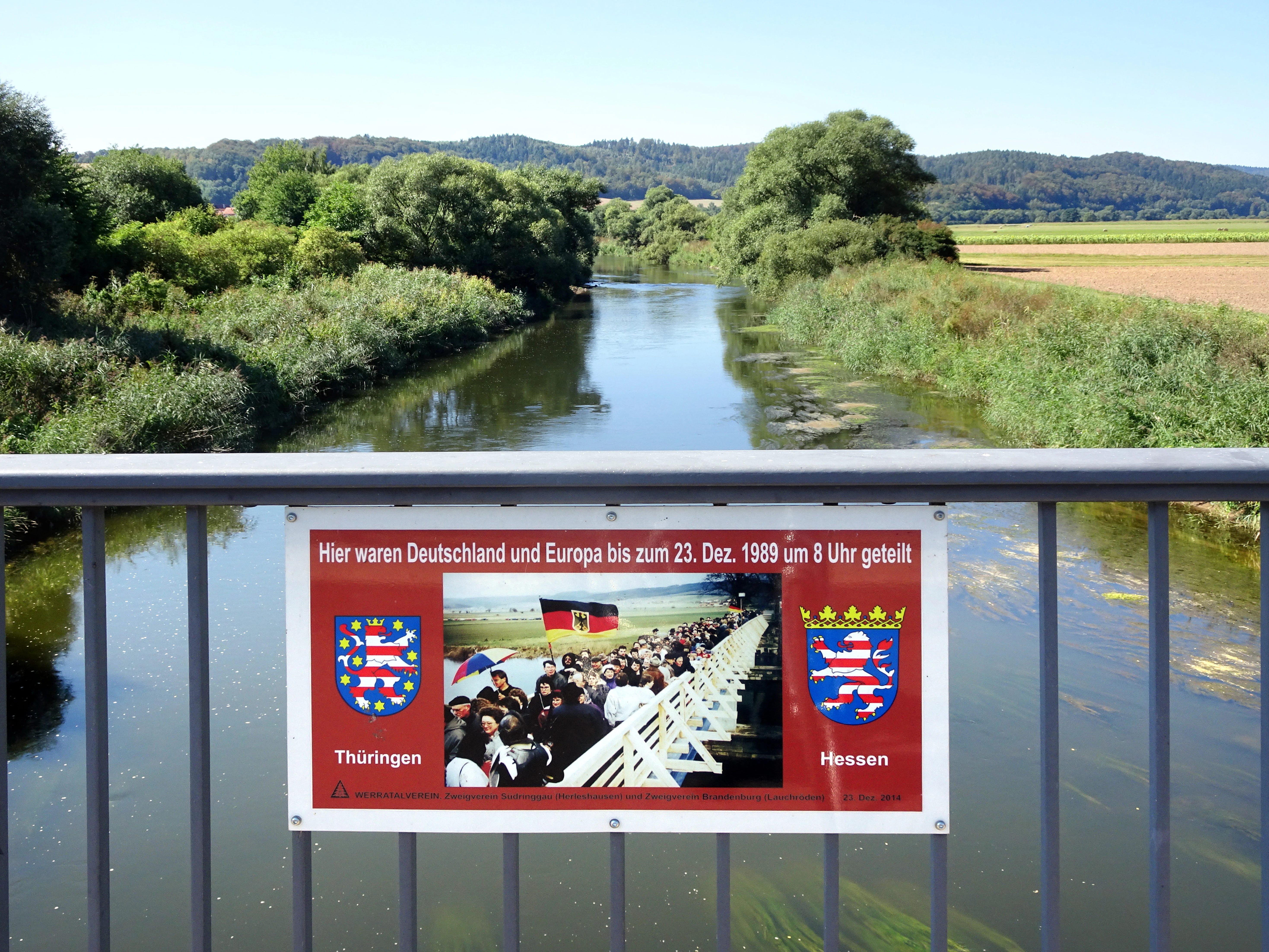 A rectangular information sign attached to a bridge railing over a river, featuring text about the 1989 German border division, historic photos, and the coats of arms for Thuringia and Hesse.