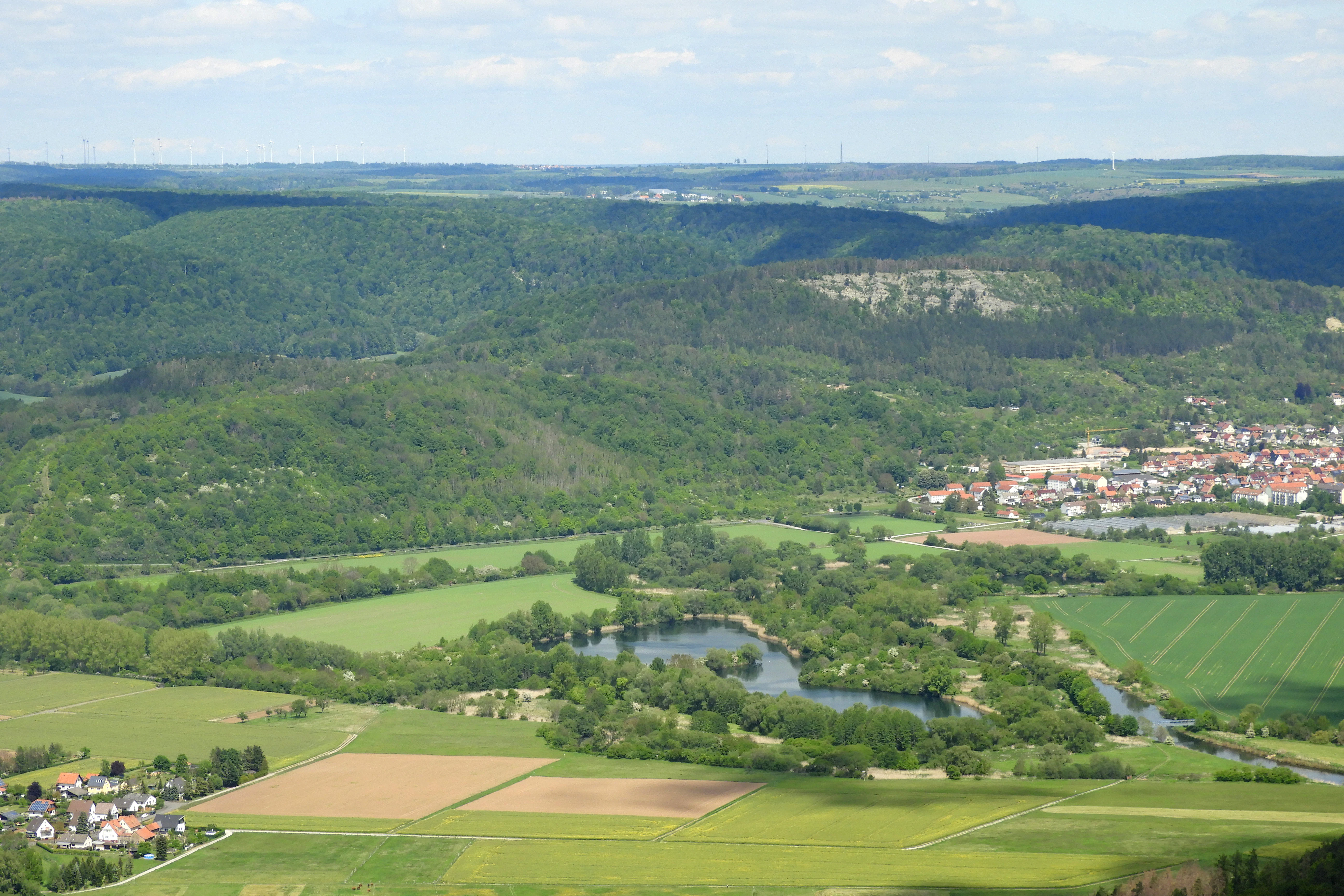 An expansive high-angle view of a river valley with a winding river, small lakes, agricultural fields, and a distant small town framed by rolling green forested hills.
