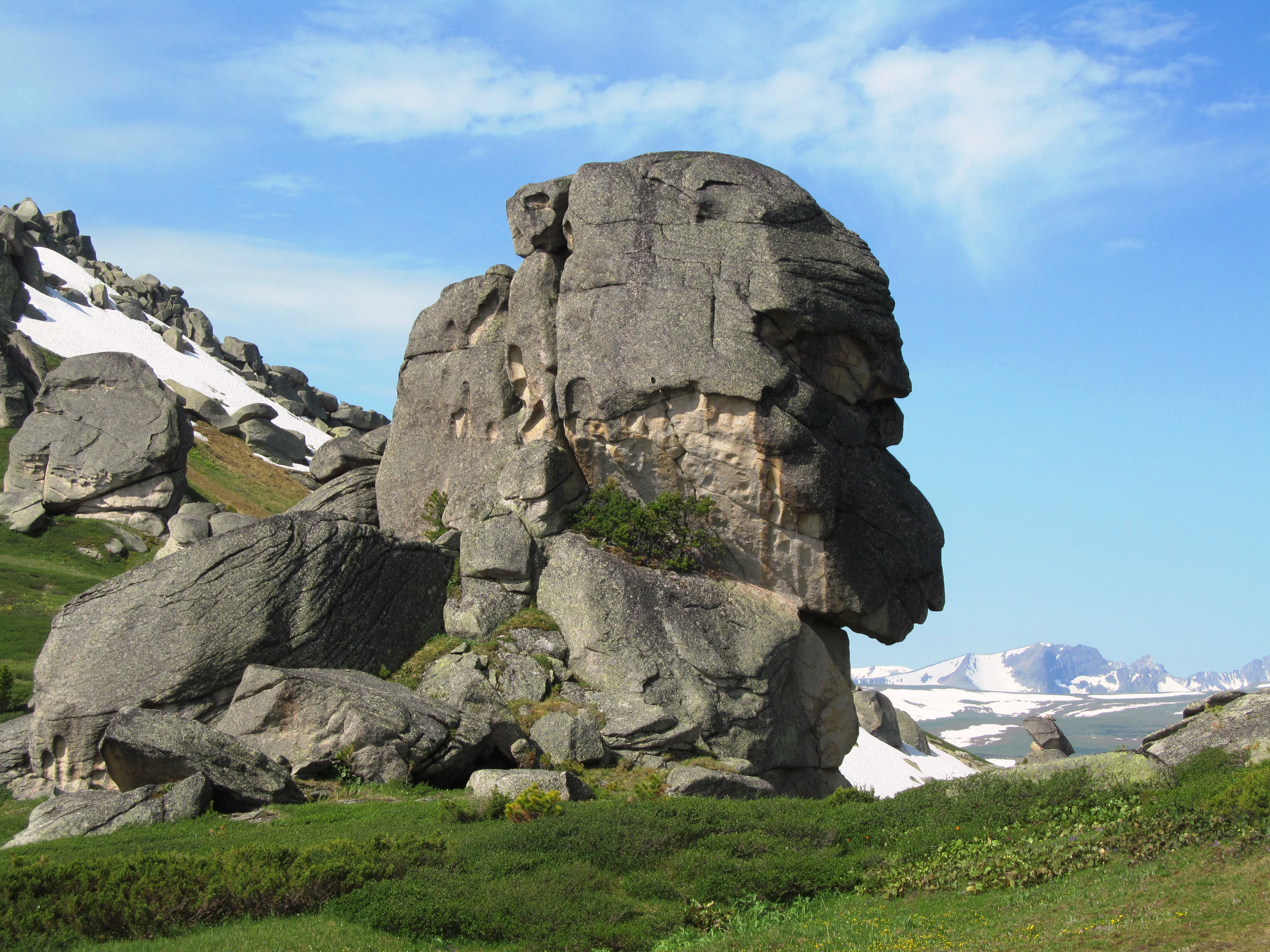 Large weathered rock formations rising from a green alpine meadow with patches of snow and a blue sky in the background.