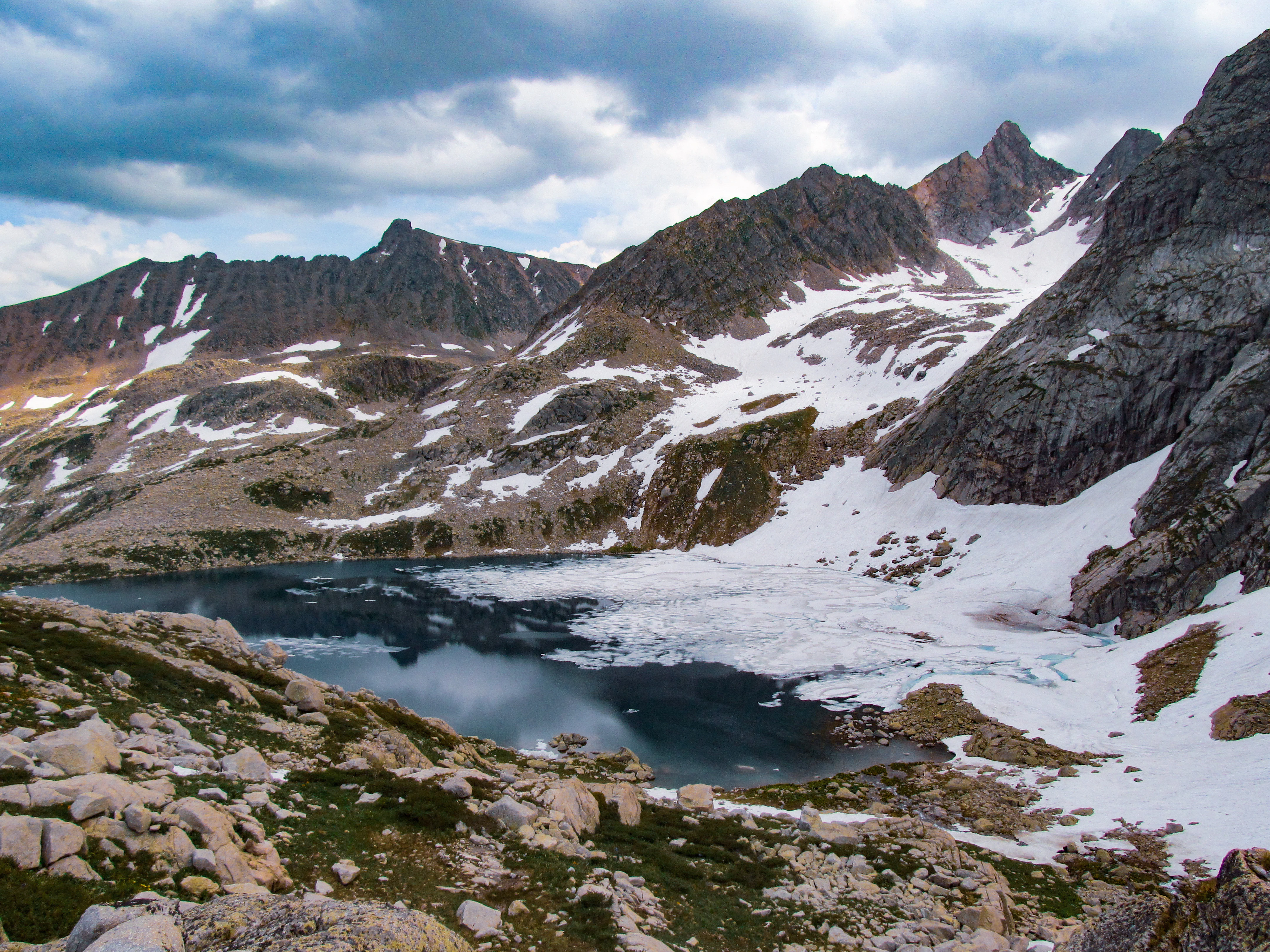 A partially frozen alpine lake surrounded by rocky mountains with snow patches under a cloudy sky.