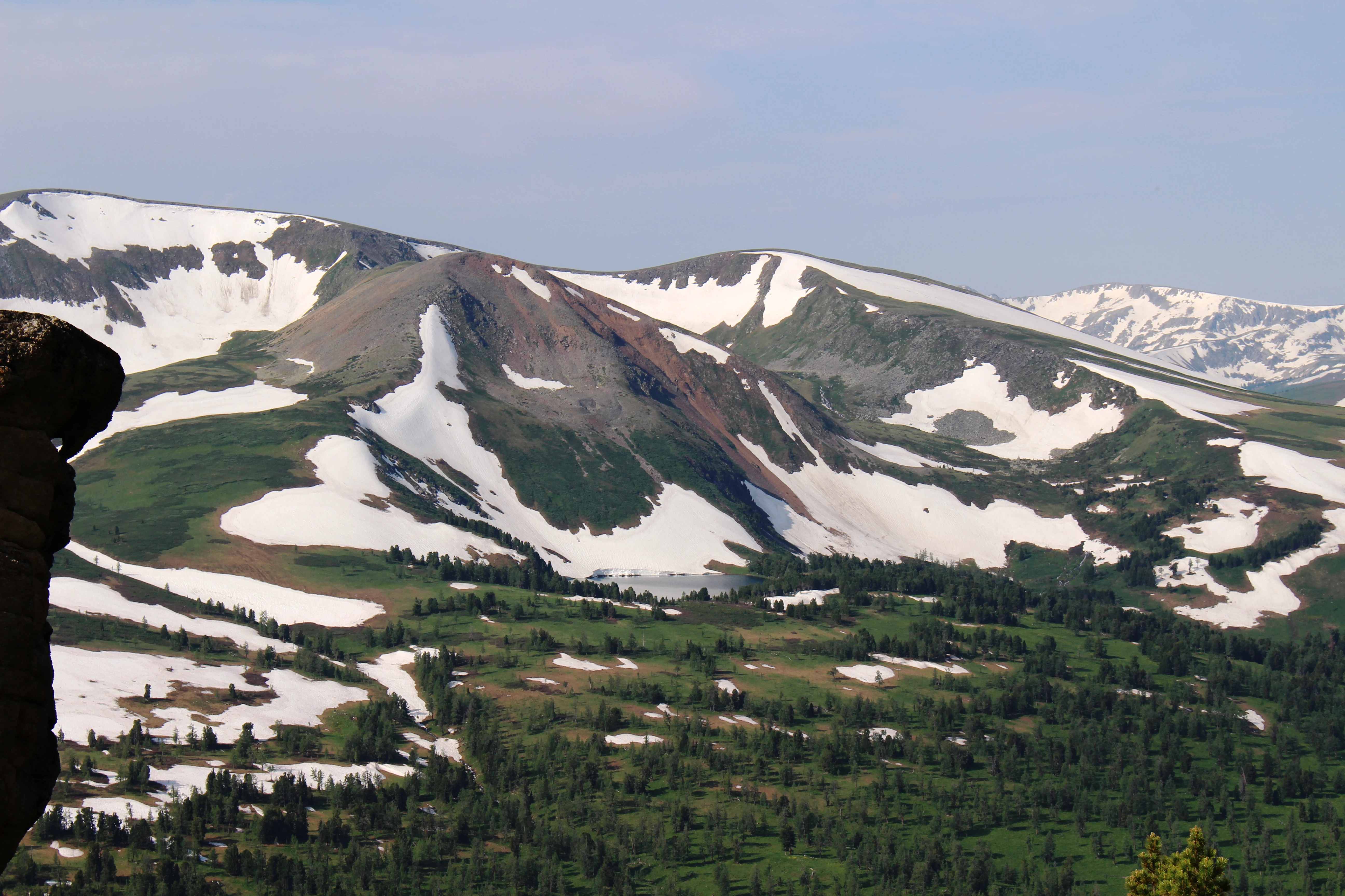 A mountain range with snow patches and green forested valleys, viewed from a rocky outcrop under a hazy sky.