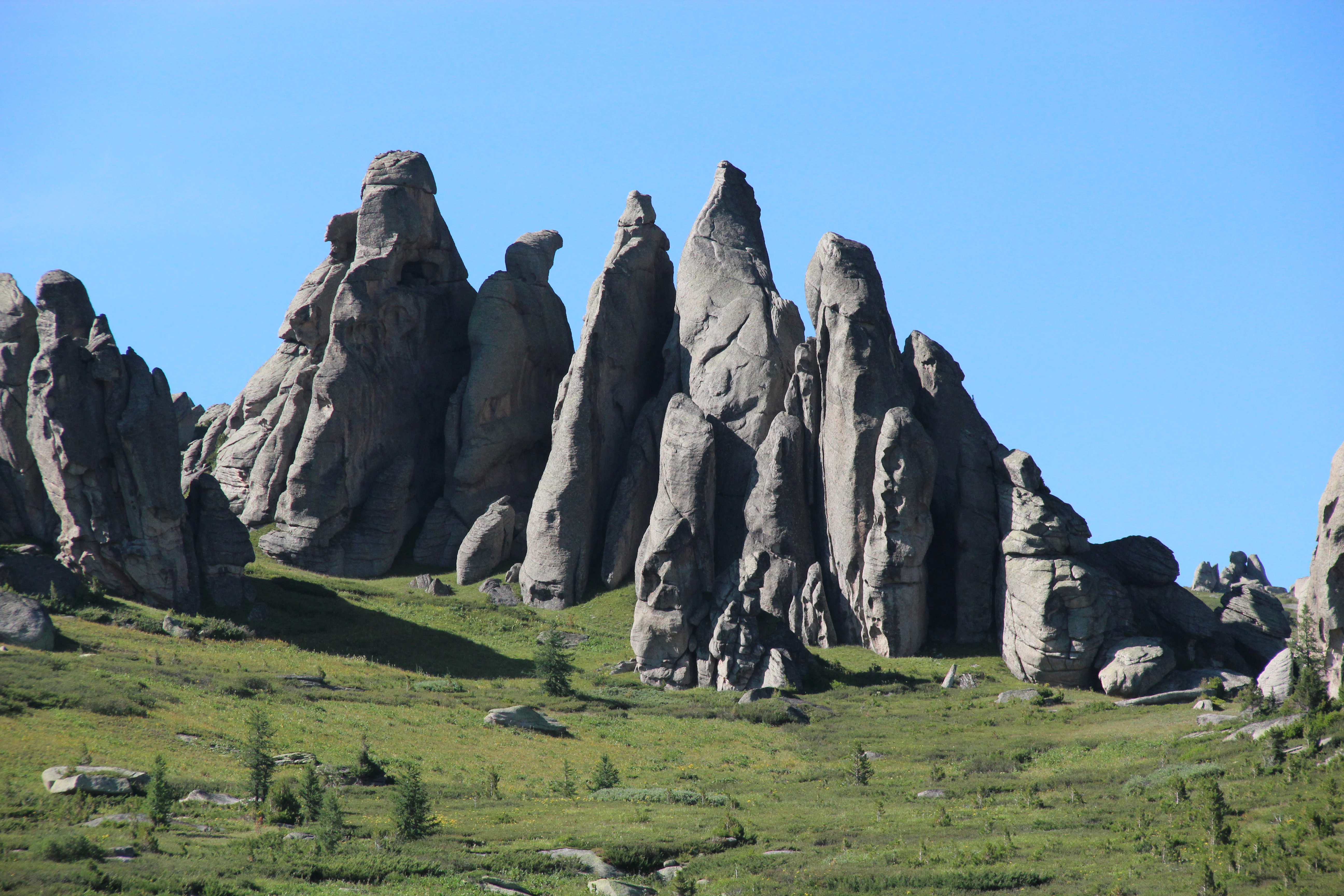 A cluster of tall vertical grey granite rock formations standing on a green grassy hill against a clear blue sky.