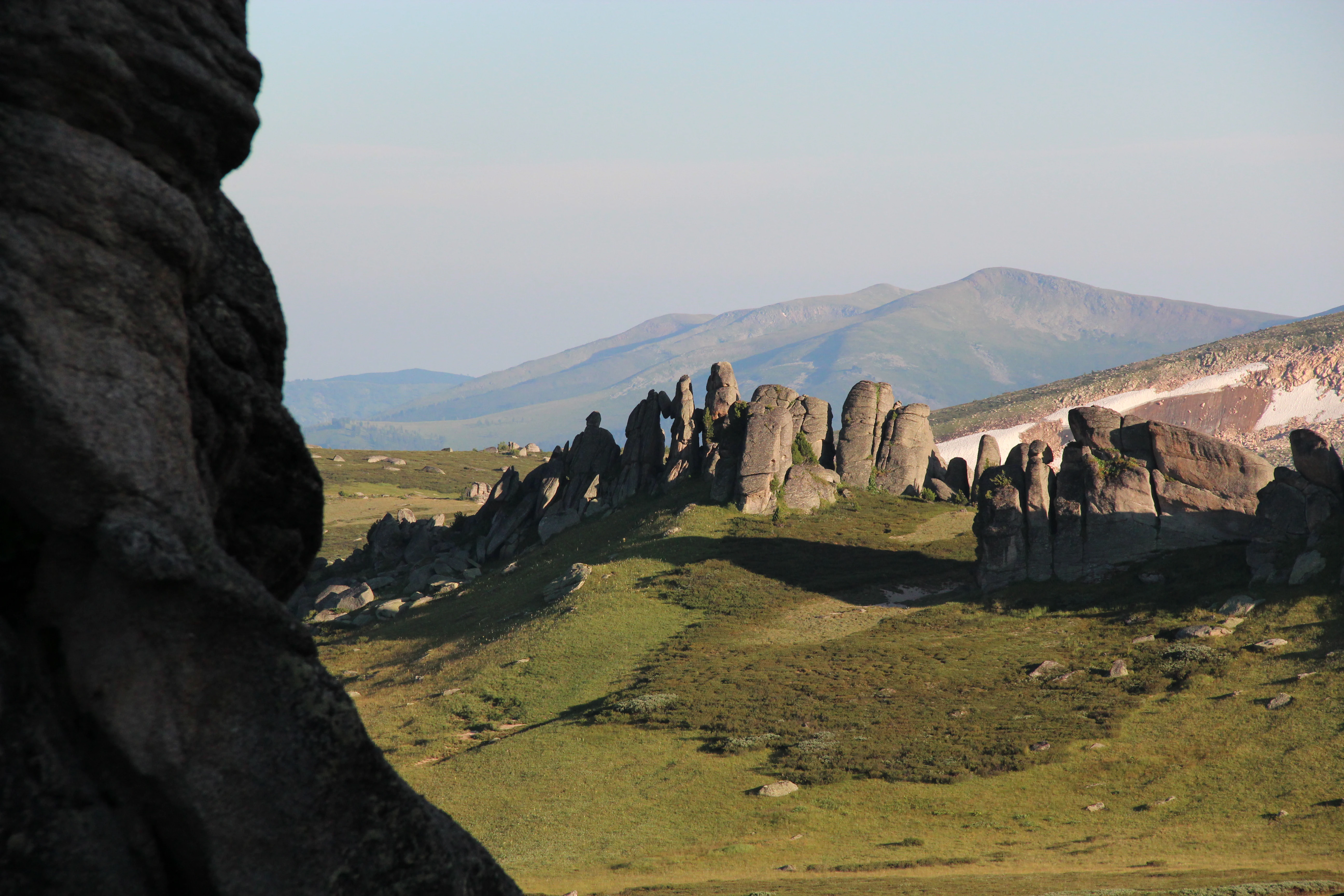 A landscape view of distinctive granite rock pillar formations rising from a green grassy hillside, with a large dark rock formation in the left foreground and rolling mountains visible in the background under a pale sky.