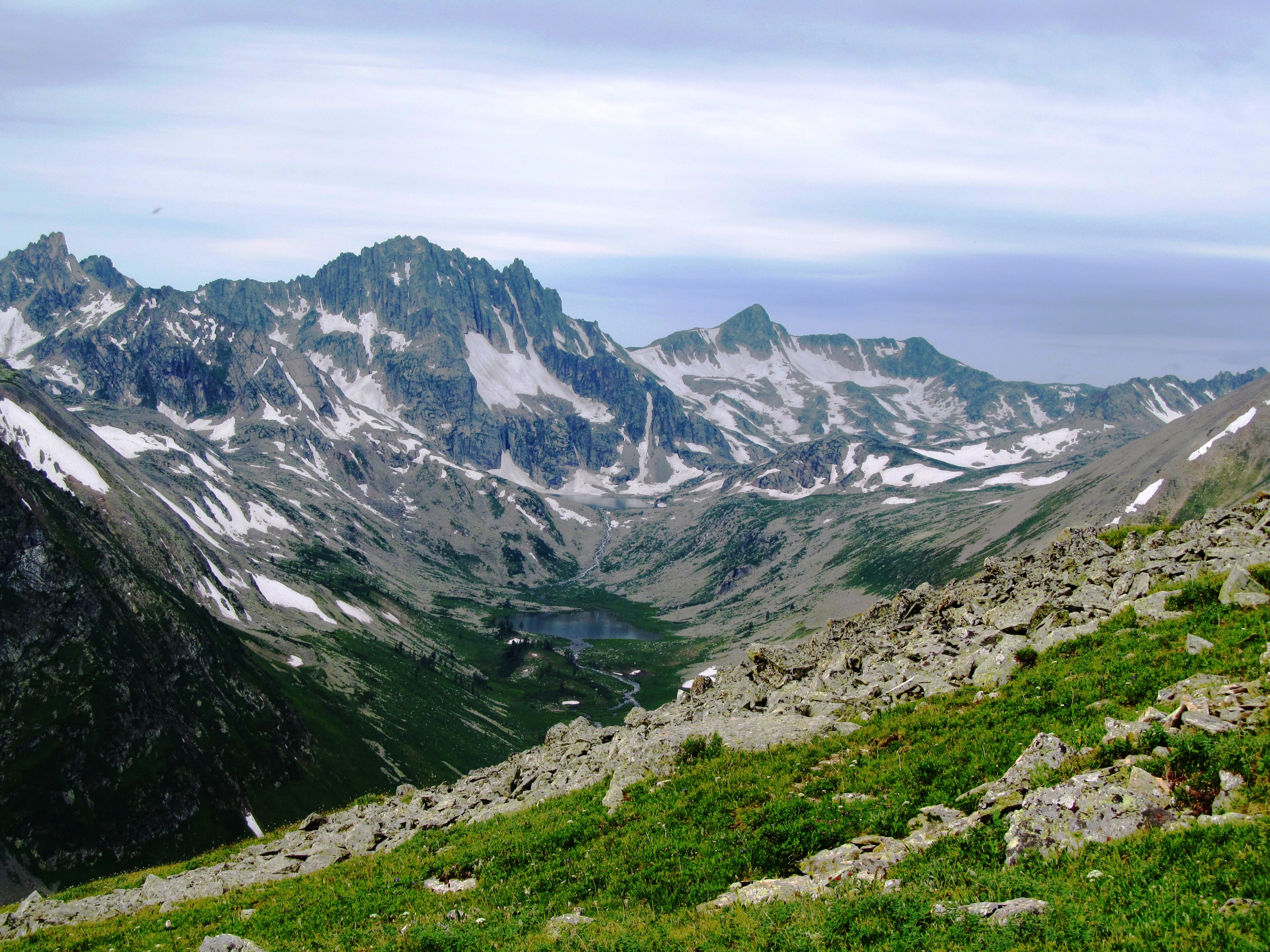 A wide mountain landscape showing rocky alpine peaks with snow patches, a green vegetated slope in the foreground, and a small lake nestled in the valley below under an overcast sky.