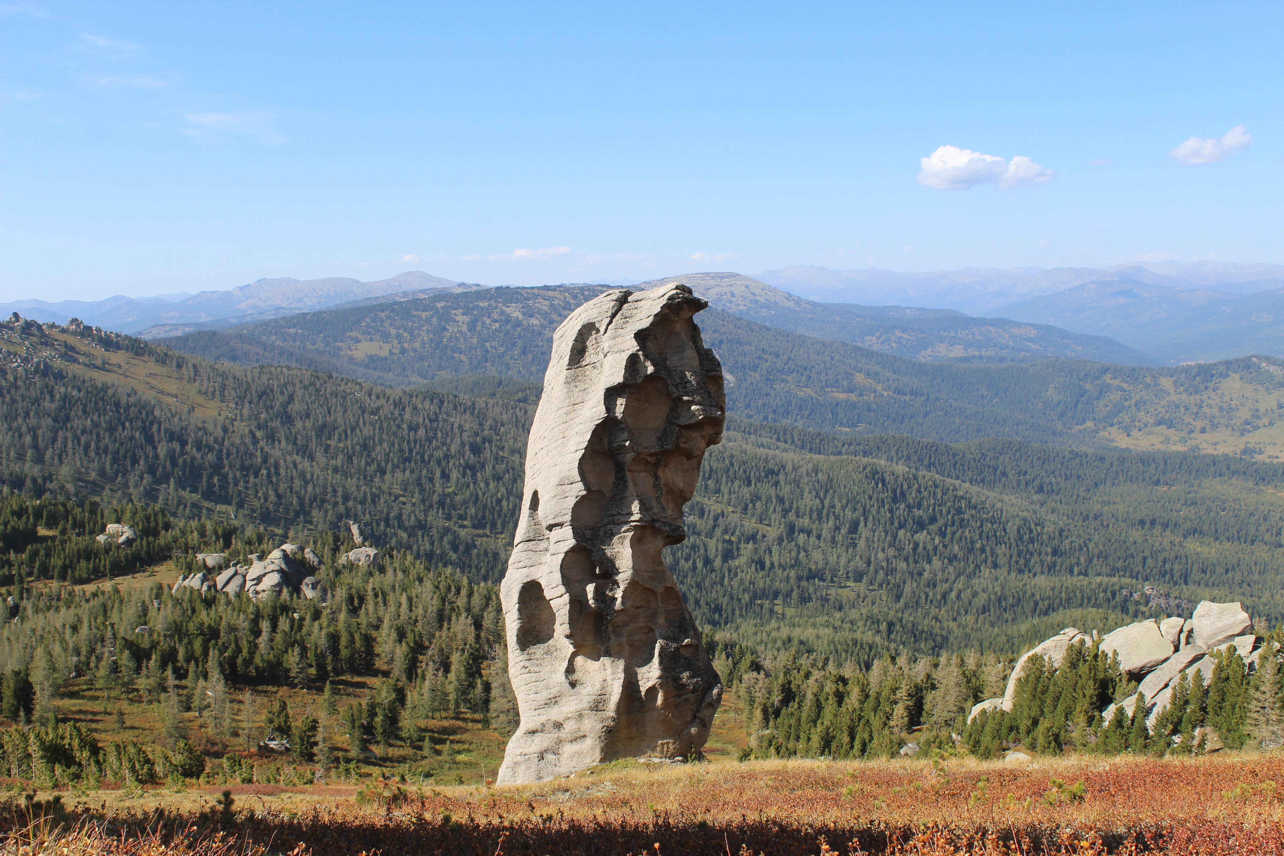 A tall, weathered granite rock pillar stands centrally in a mountainous landscape covered in green coniferous forest under a blue sky with scattered clouds.