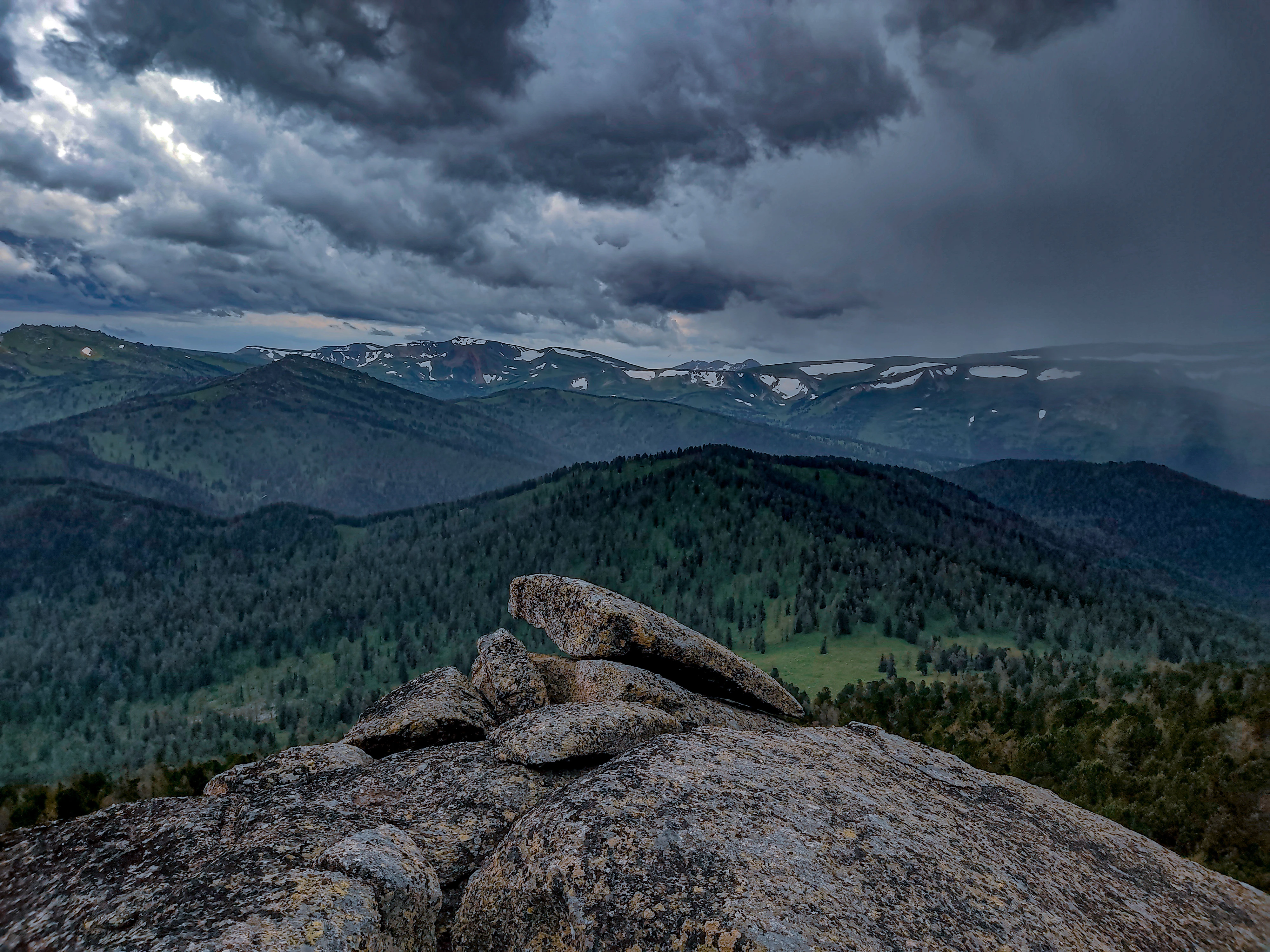 A rocky foreground with a large boulder, overlooking a vast valley of green coniferous forest and distant mountains with patches of snow under a dark, cloudy sky.