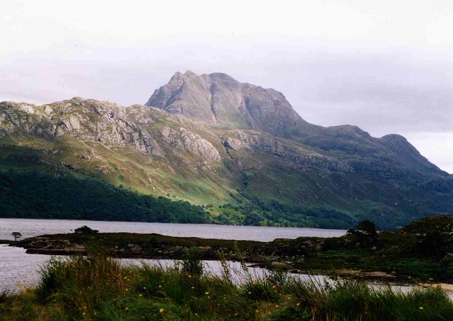 A wide landscape view of a large, rugged mountain peak, Slioch, looming over the calm, dark surface of Loch Maree, with green slopes and a rocky, vegetated foreground.