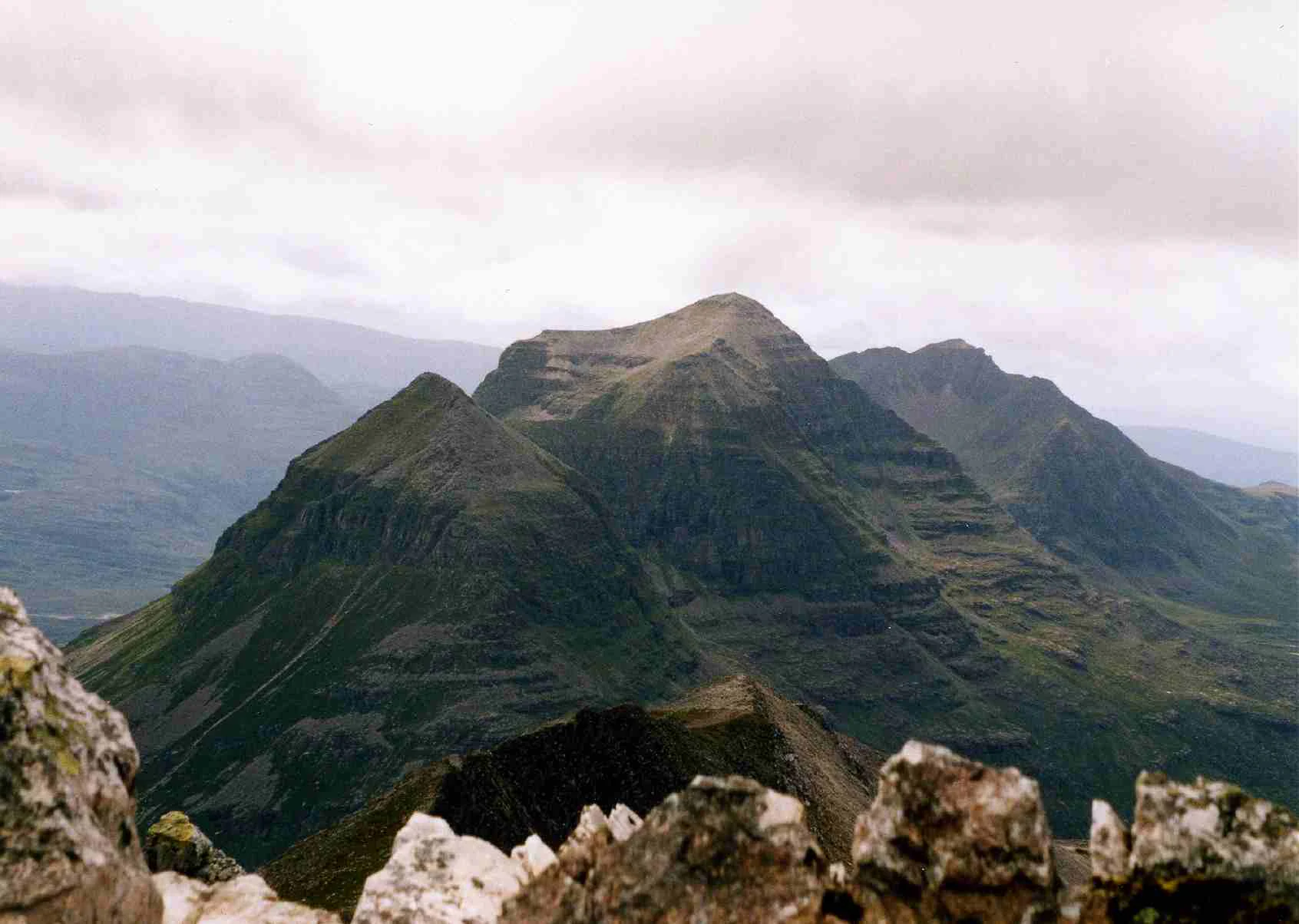 A wide shot of a jagged mountain massif with steep, terraced slopes under a cloudy sky, seen from a rocky high-altitude vantage point.