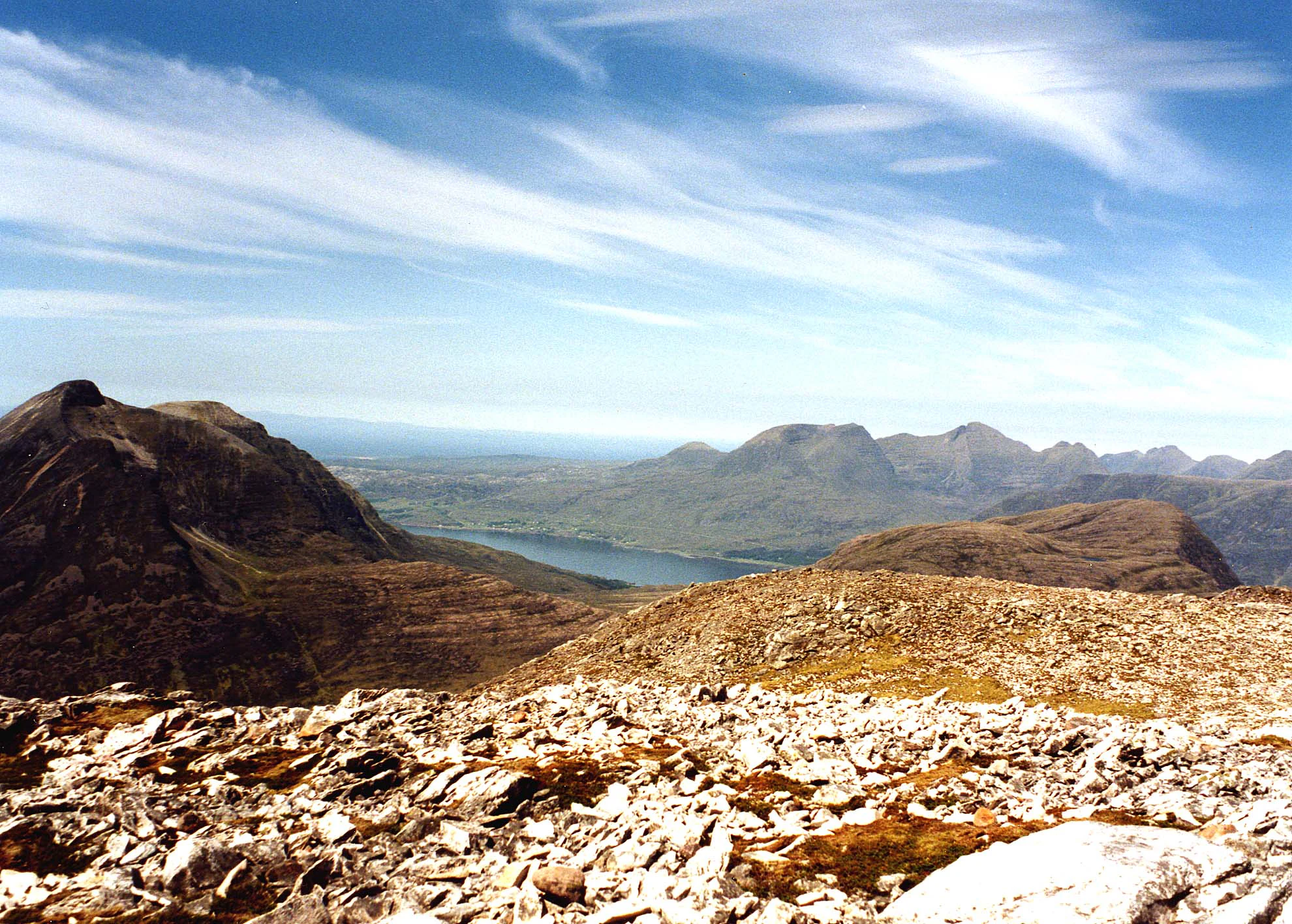 A high-angle view looking over a rocky, desolate mountain summit toward a deep valley with a dark lake and layered mountain ridges under a blue sky with wispy clouds.