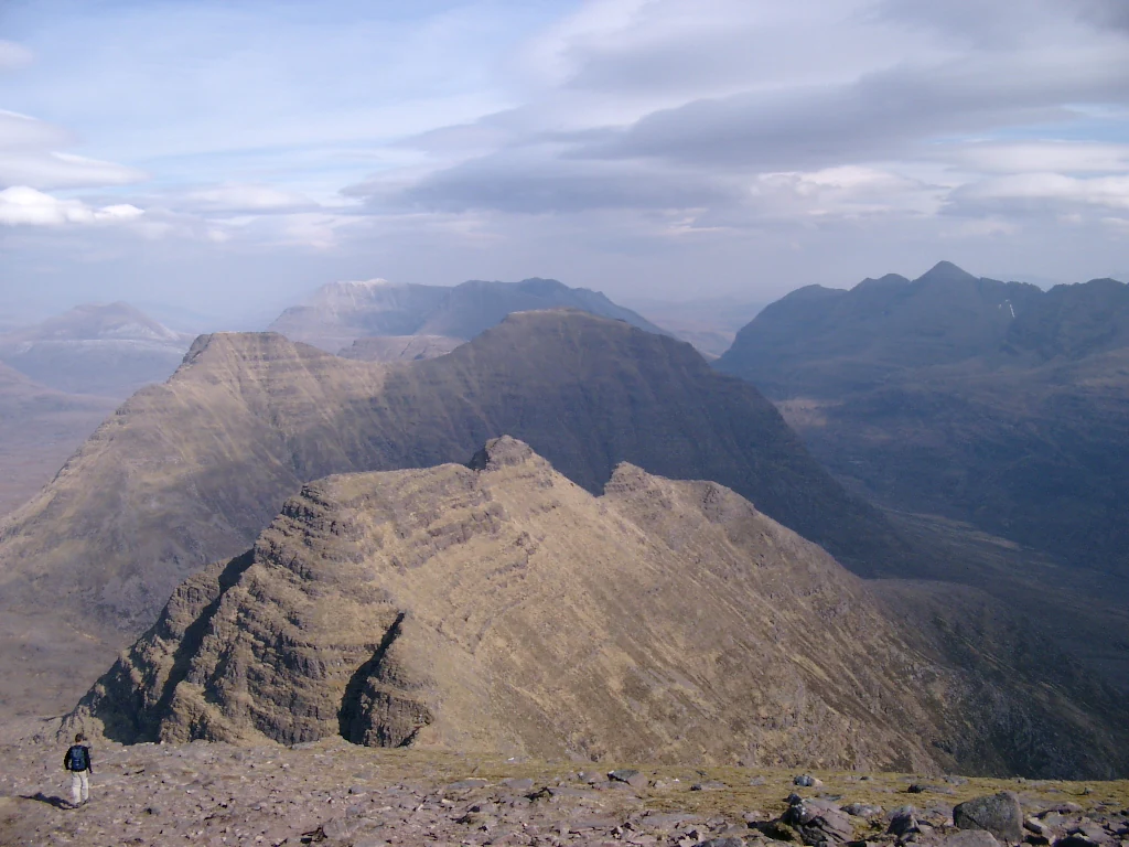 A wide view of jagged mountain peaks under a cloudy sky in the Scottish Highlands, with a lone hiker in the bottom left foreground.