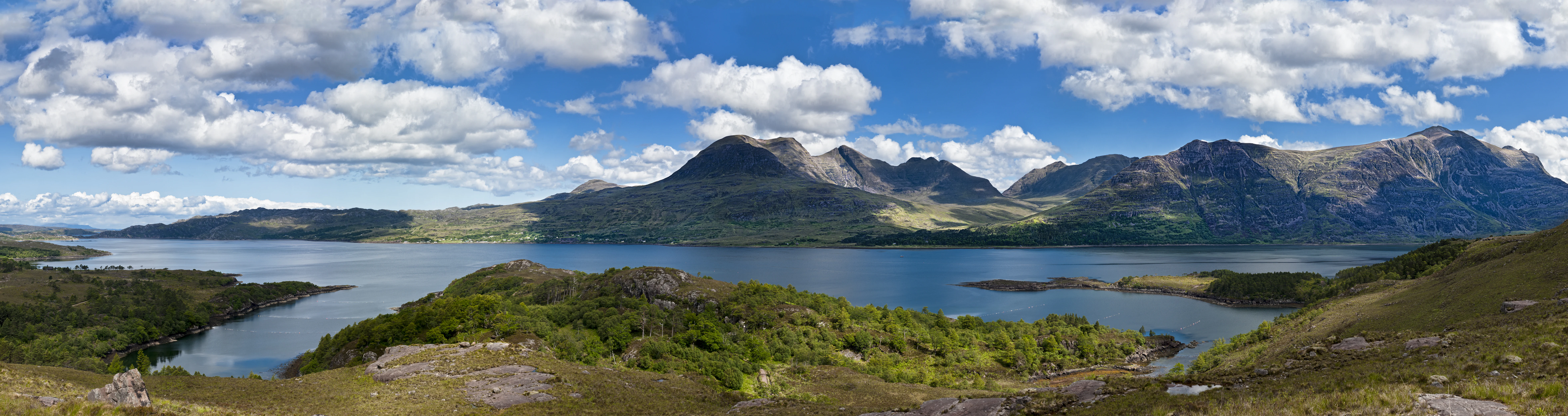 Panoramic view of a calm, large sea loch surrounded by rolling green hills, dense forested slopes, and prominent craggy mountain peaks under a bright blue sky with scattered white clouds.