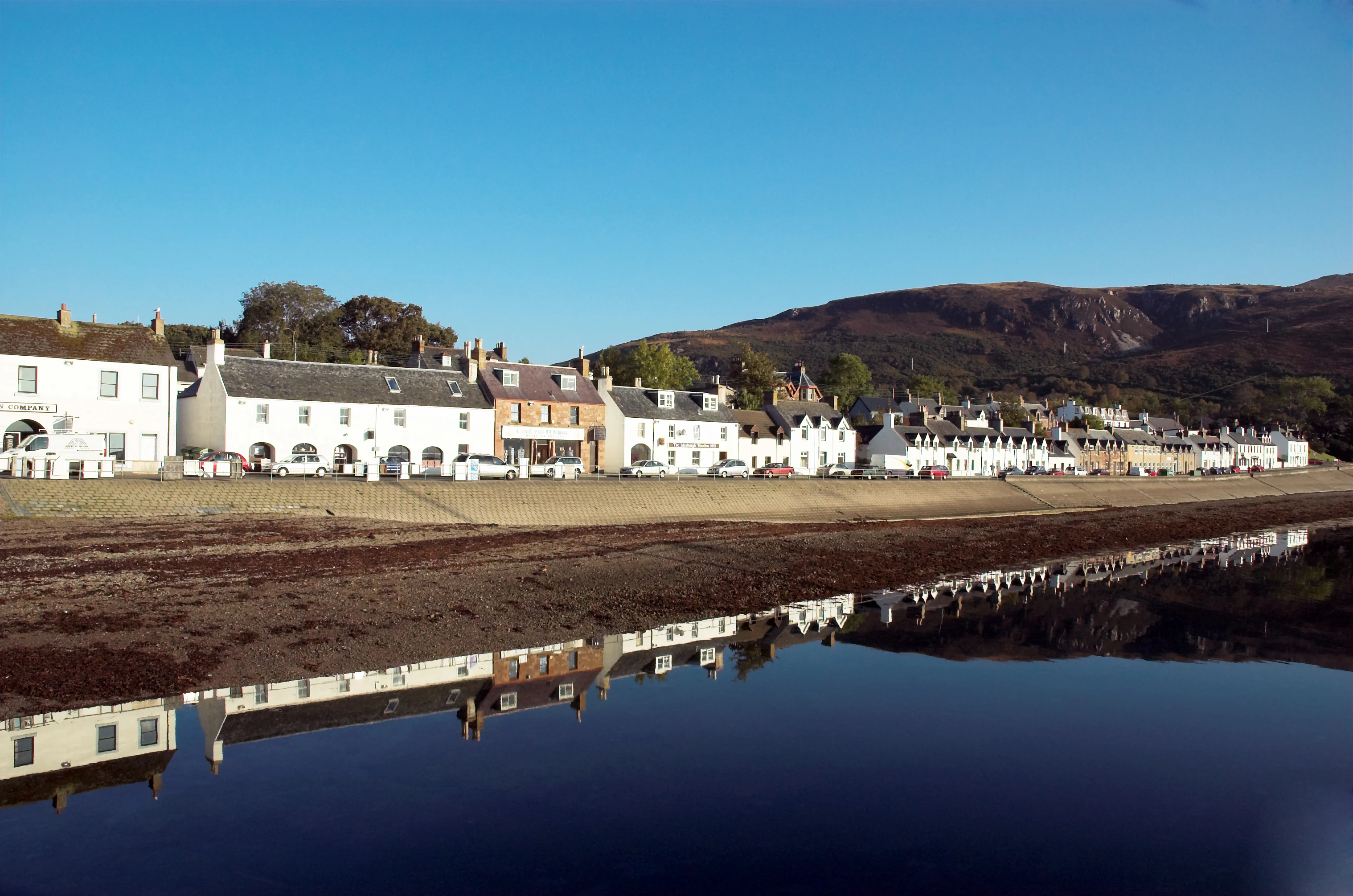A wide view of a row of white and stone buildings along a shoreline, with their reflections visible in calm water and hills in the background under a clear sky.