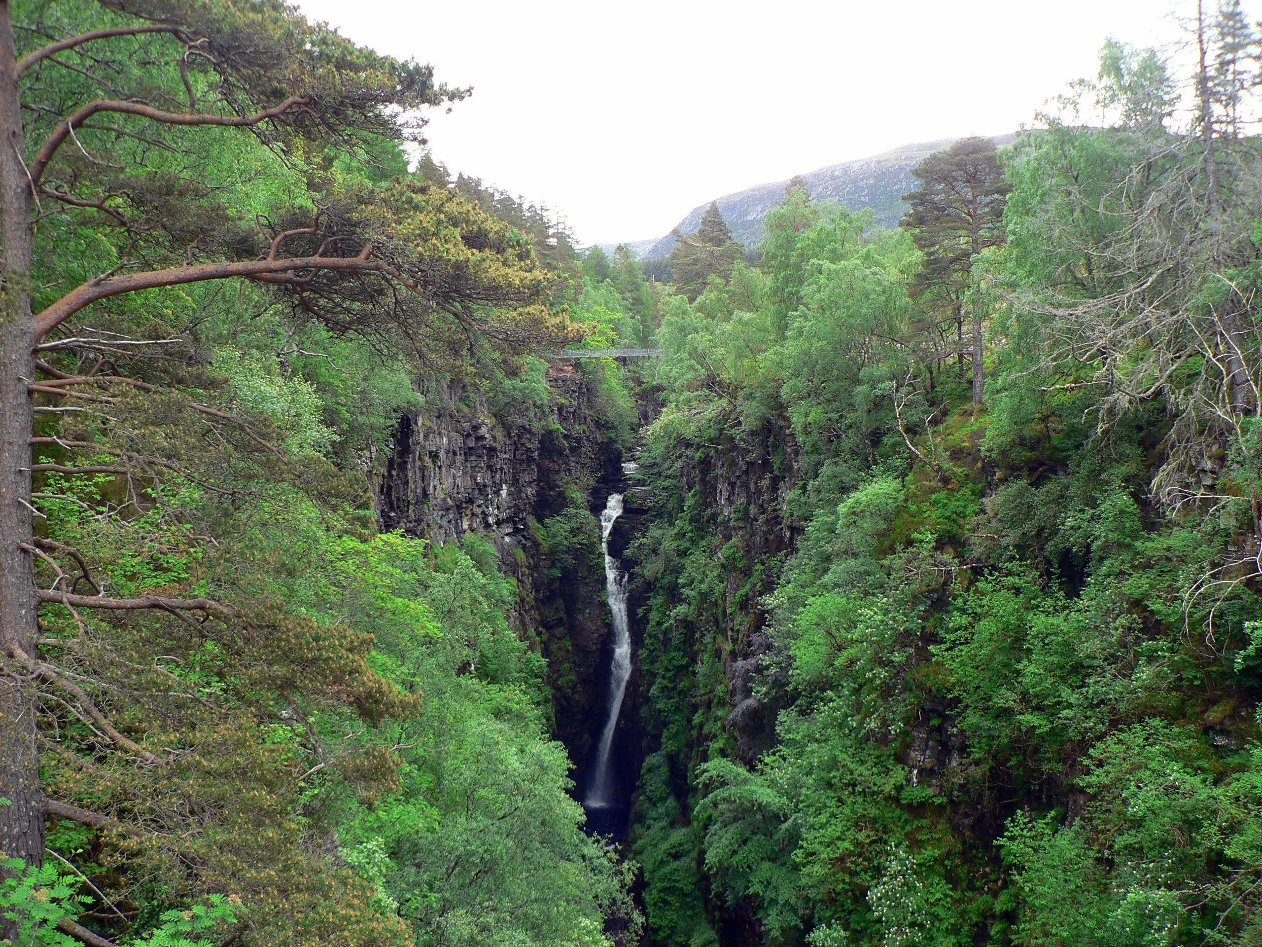 A narrow, deep gorge with a waterfall cascading down the center, surrounded by lush green forest and rocky cliffs.
