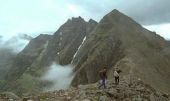 Two hikers walking along a rocky, steep mountain ridge with a towering, sharp-peaked mountain massif in the background under an overcast sky.