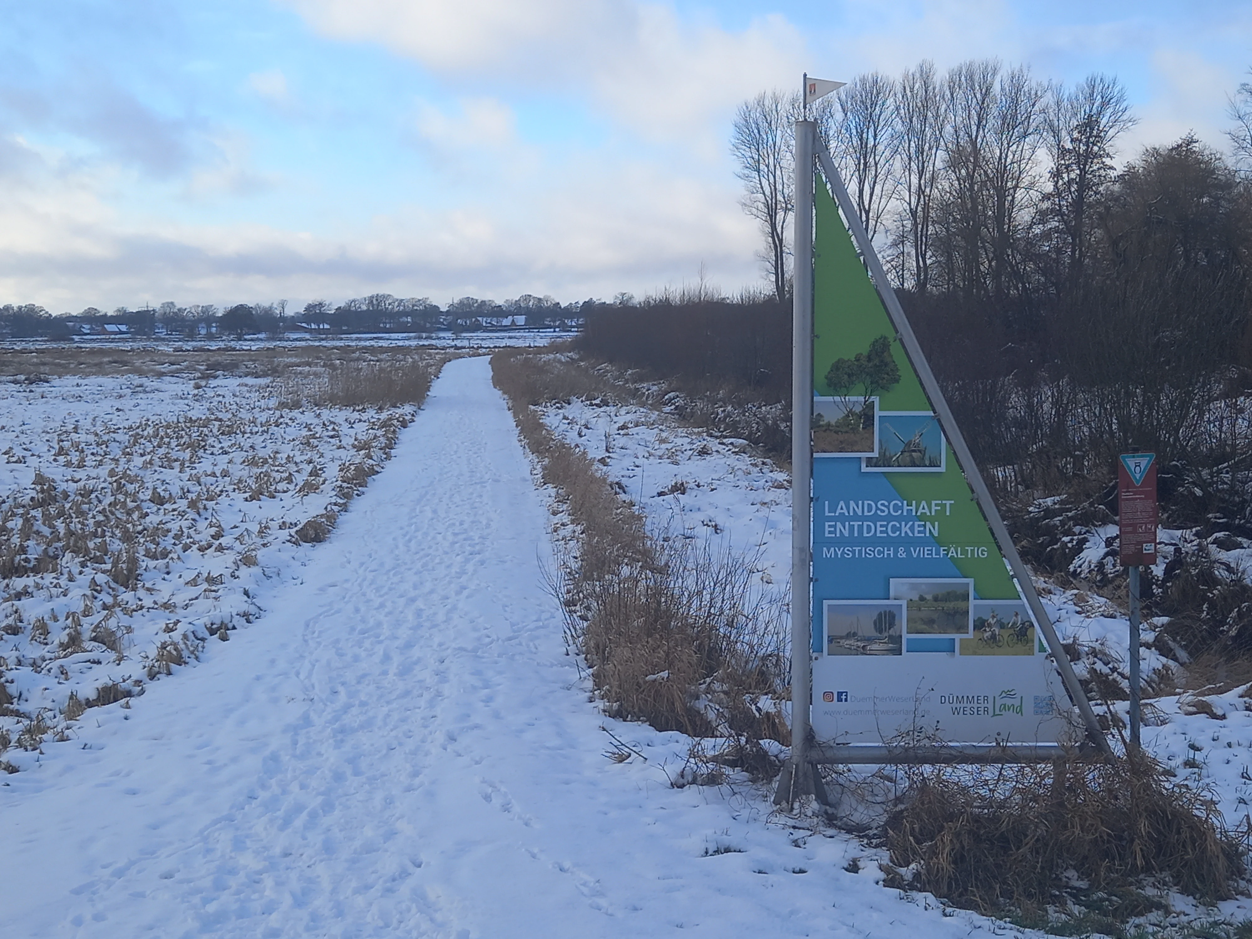 A snow-covered trail winds through a winter landscape, with a triangular informational signpost in the foreground reading Landschaft entdecken.