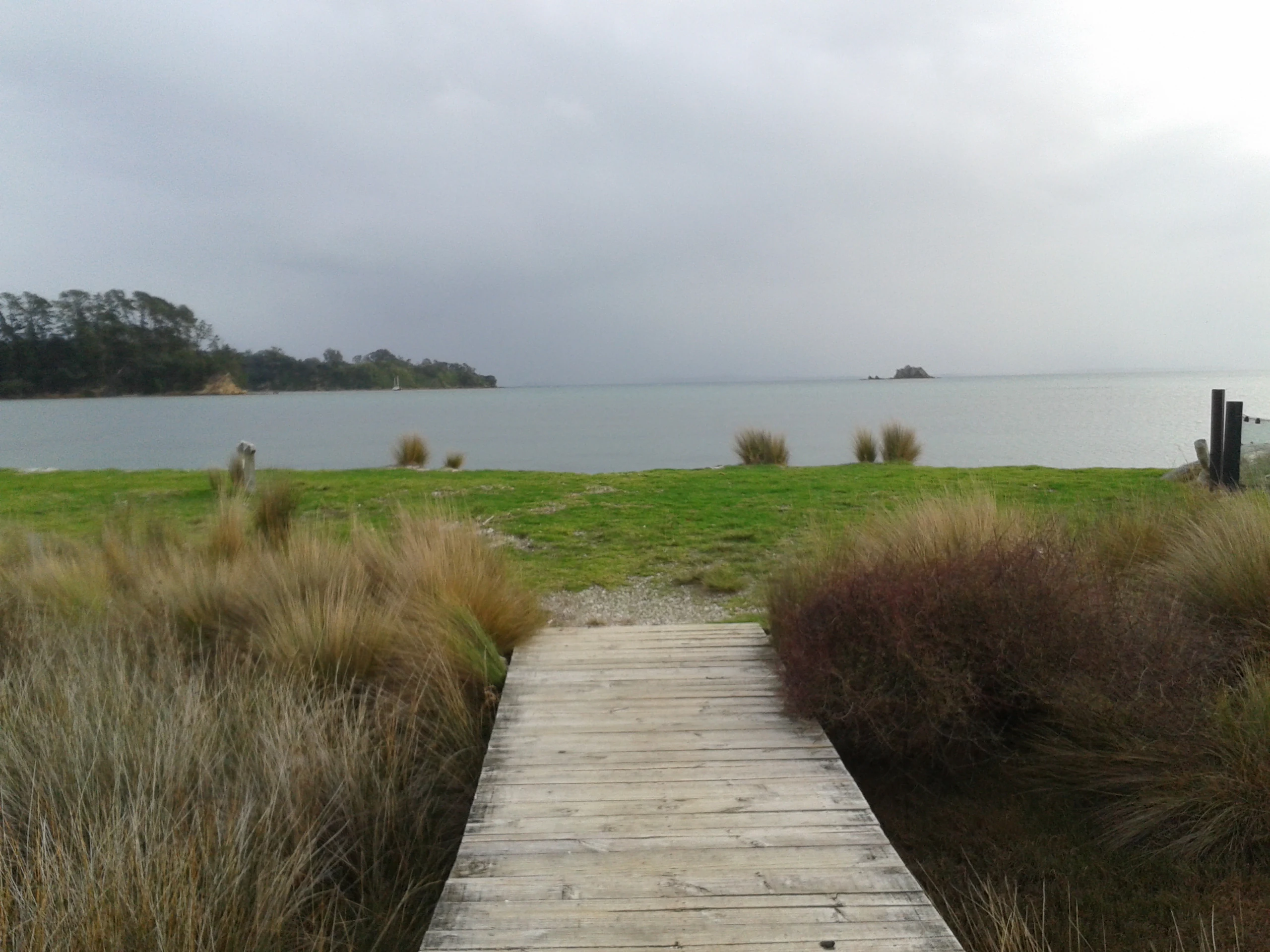 A wooden boardwalk path leading through grasses to a calm bay under an overcast sky at Whakanewha Regional Park.