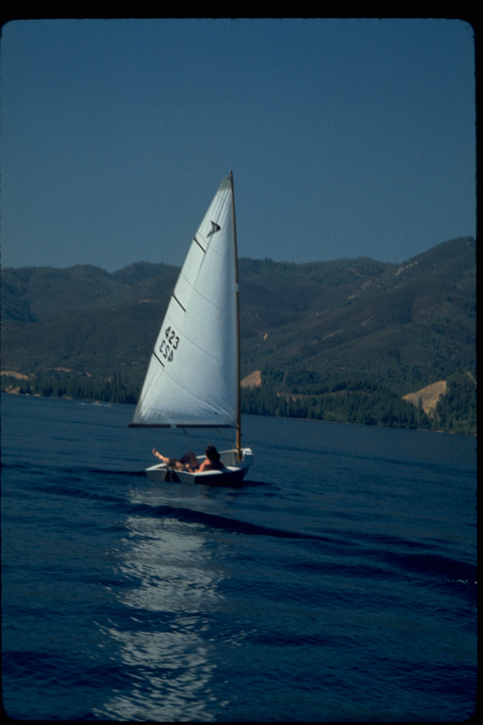 A person sailing a small white sailboat on a calm blue lake with forested mountains in the background.