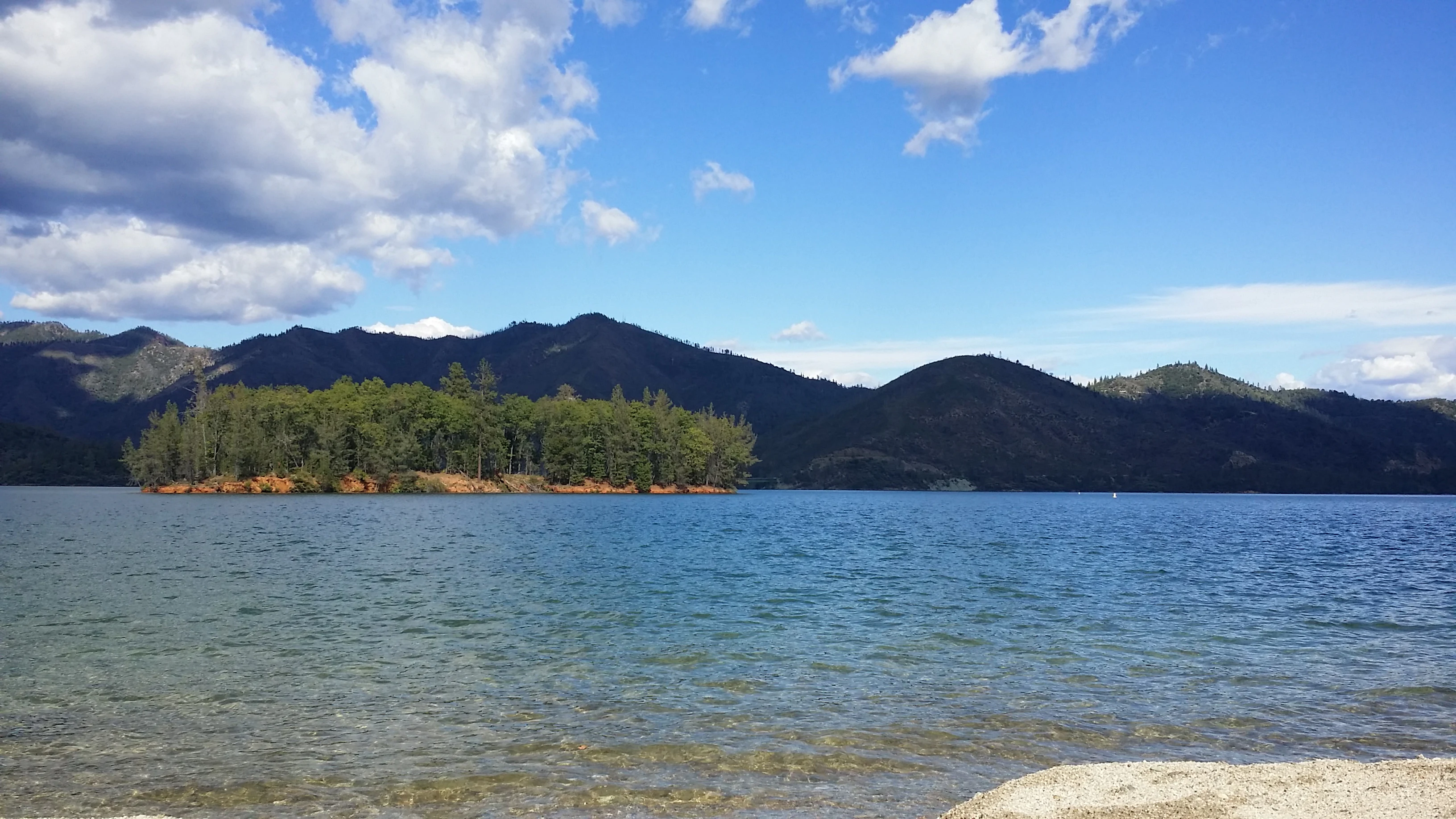 A wide angle shot of a calm blue lake with a small tree-covered island in the center, framed by distant forested mountains under a blue sky with scattered white clouds.