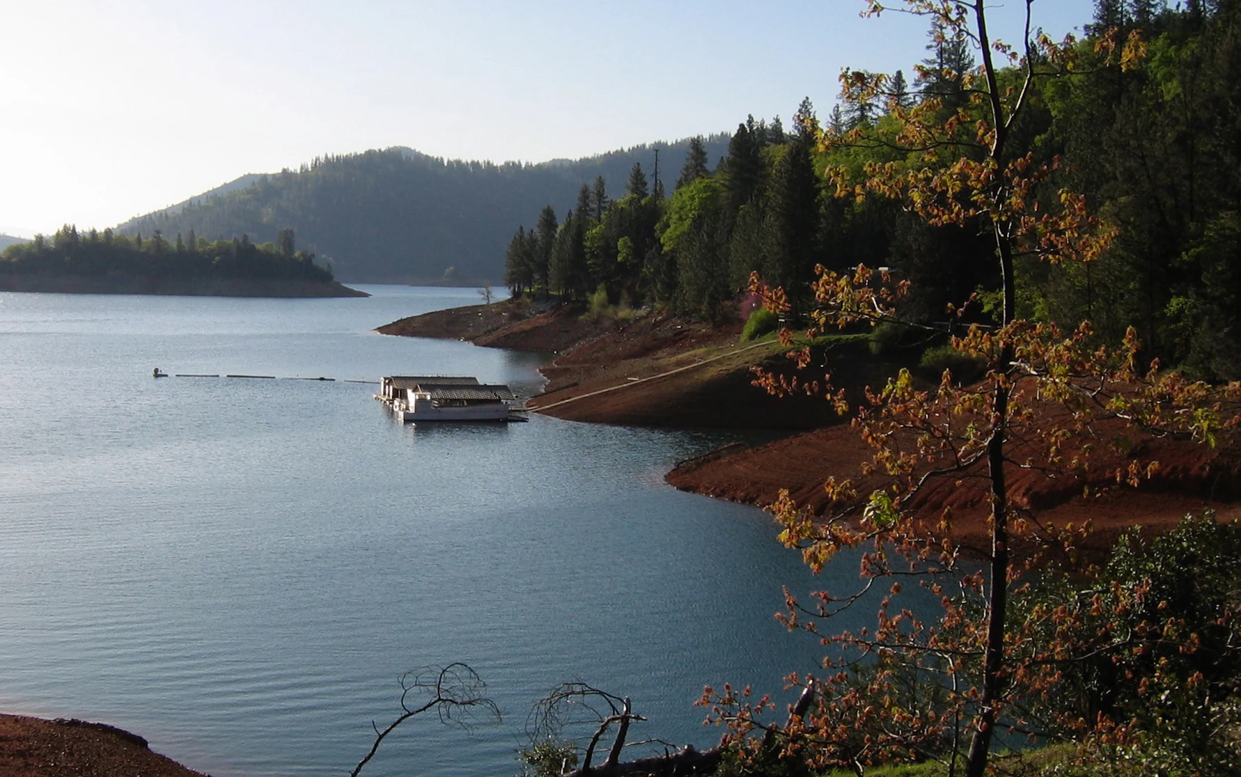 A wide angle view of a reservoir, Shasta Lake, with a floating dock on the water, a reddish-brown shoreline, and forested hills in the background under soft sunlight.