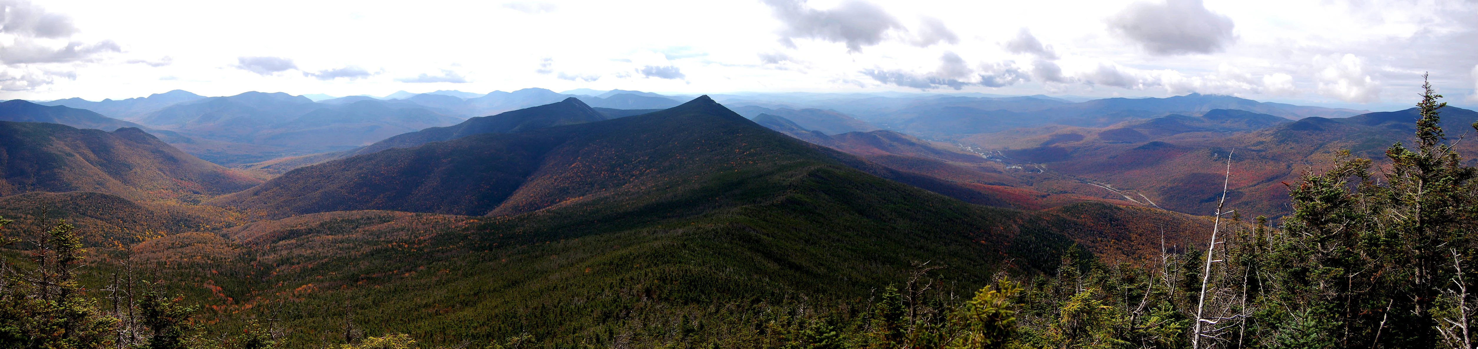 A sweeping panoramic view of forested mountain ranges under a partly cloudy sky, with hints of autumn foliage in the valleys.
