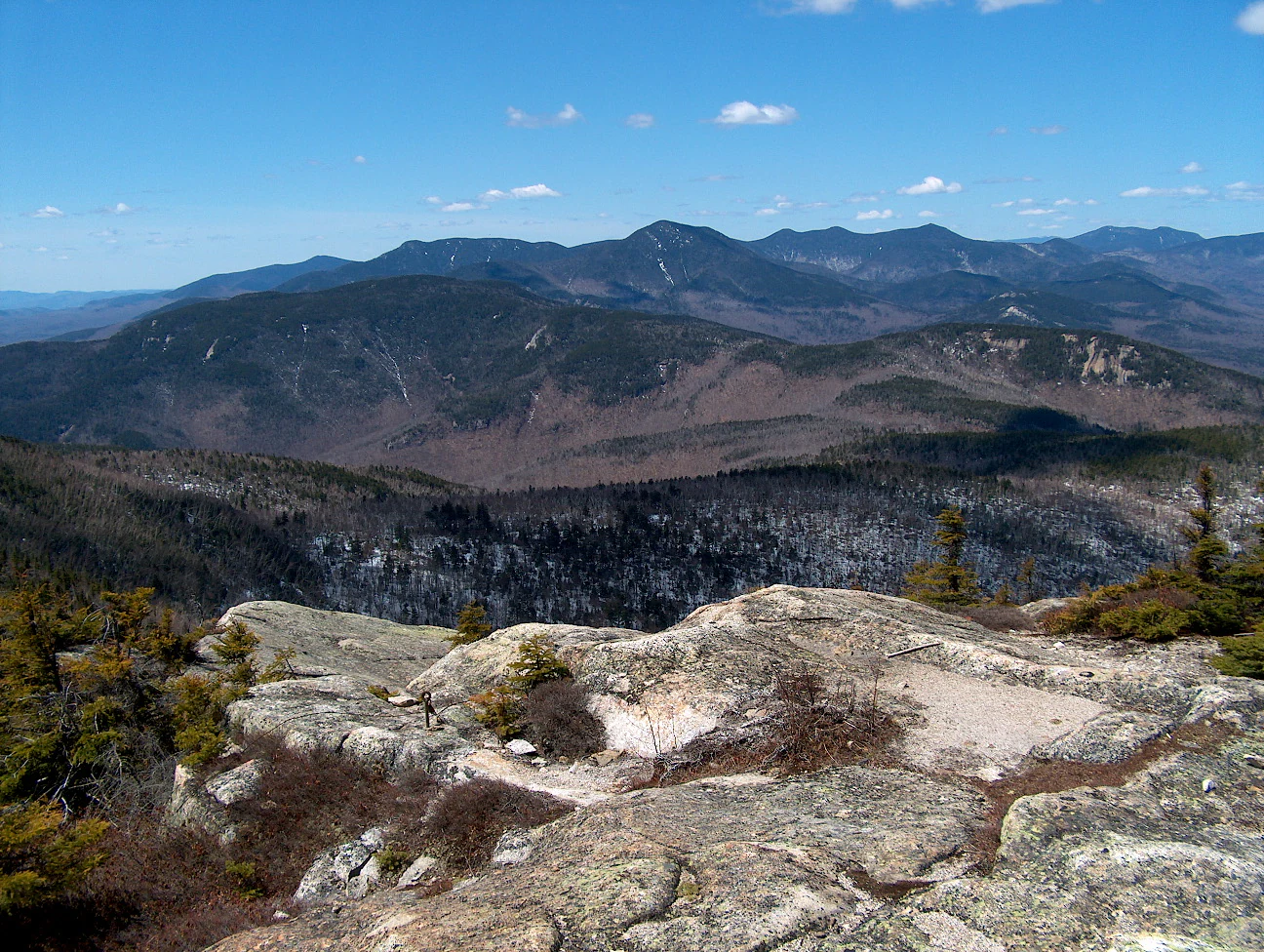 A high-angle, wide-angle shot looking out over the layered, deep-blue peaks of the Sandwich Range in New Hampshire, with a rocky, lichen-covered foreground and a clear blue sky above.