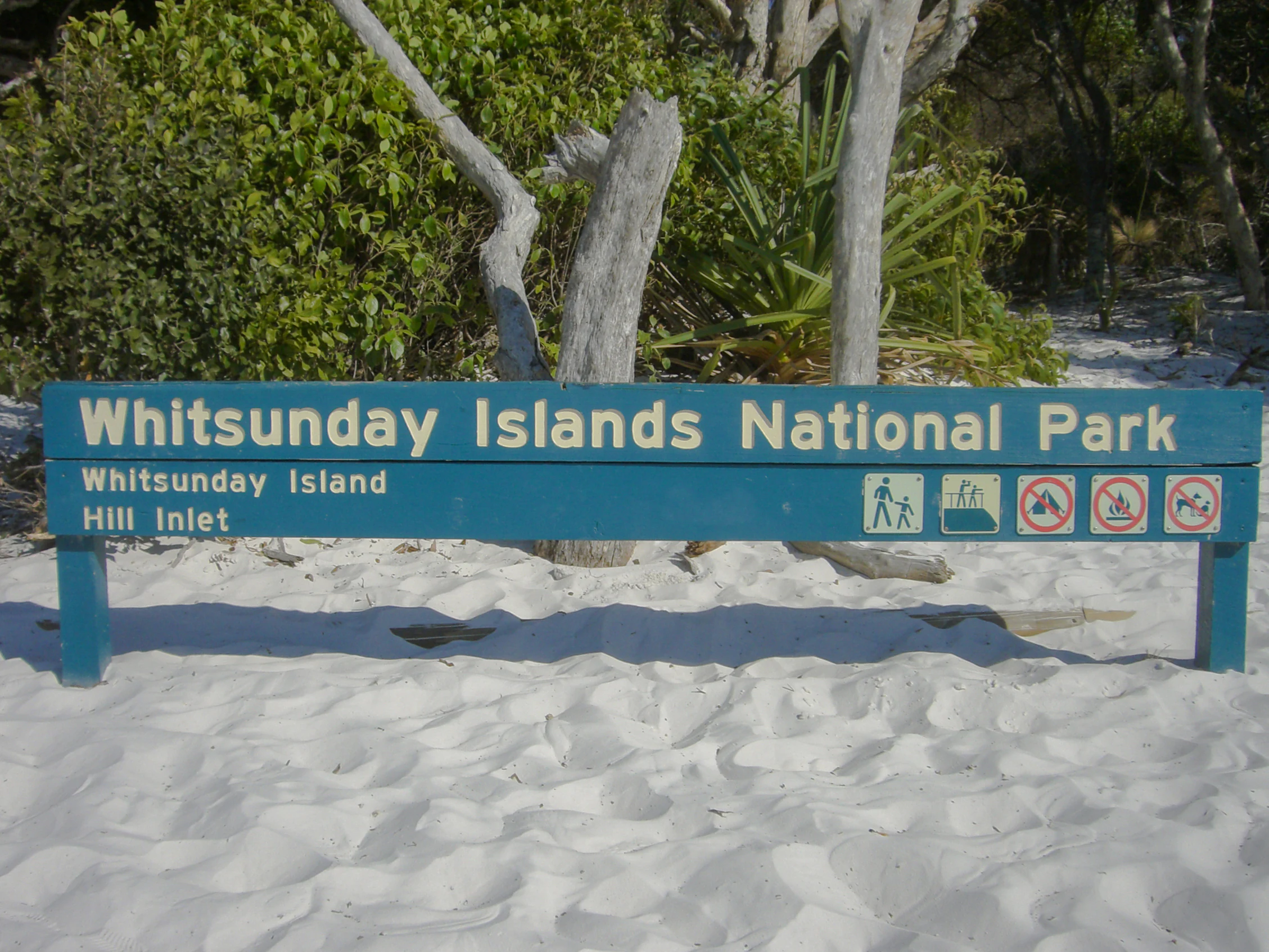 Cartel azul del Parque Nacional Islas Whitsunday con texto e iconos blancos, ubicado en una playa de arena blanca con árboles y vegetación al fondo