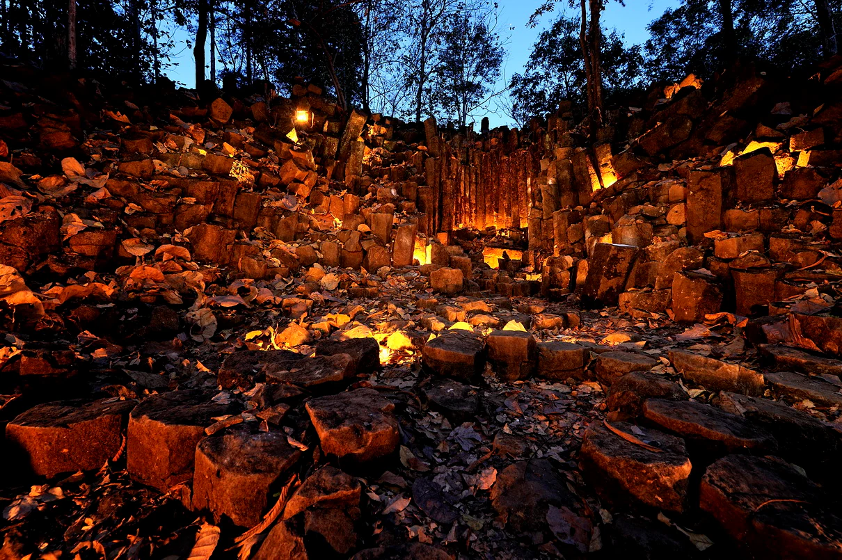 Dark landscape of columnar basalt formations with artificial lighting, surrounded by trees and a darkening sky