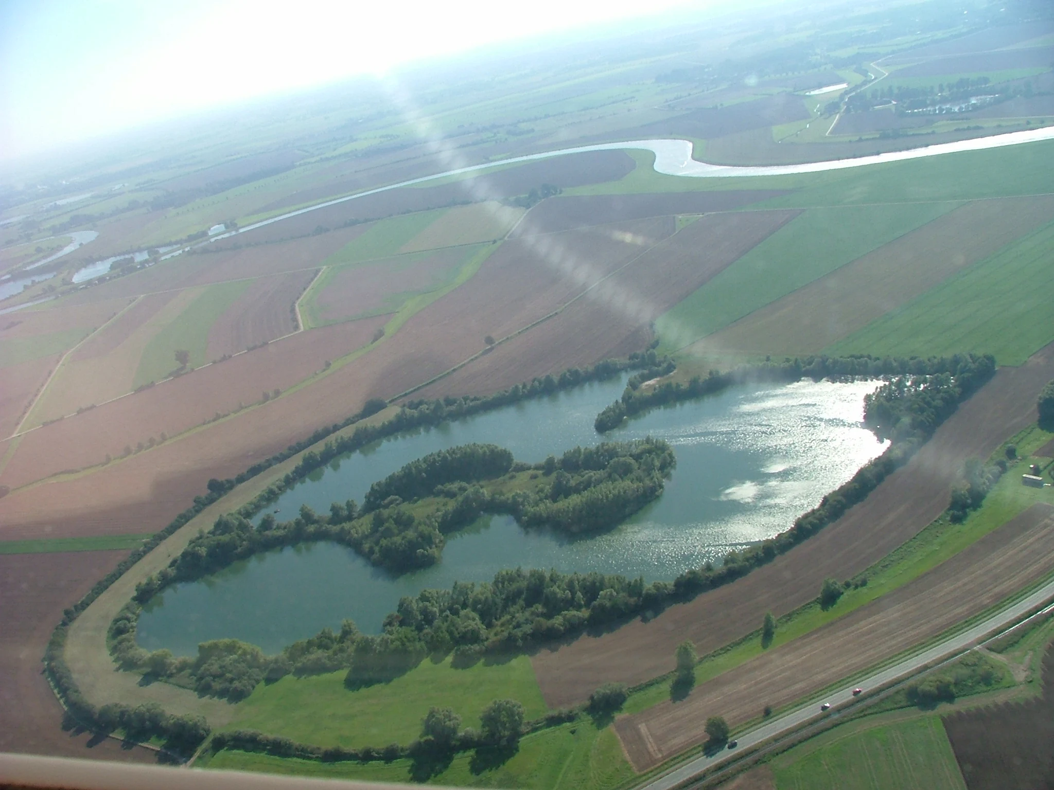 An aerial view of the Wiedesee, an elongated lake with wooded islands, surrounded by farmland and a nearby river.